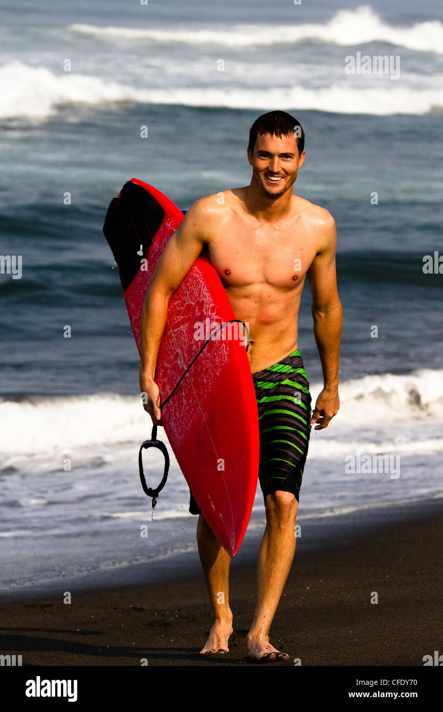 Un homme portant des shorts de surf board promenades sur la plage, à Pasquales, Mexique tout en tenant une planche rouge sous son bras. Banque D'Images