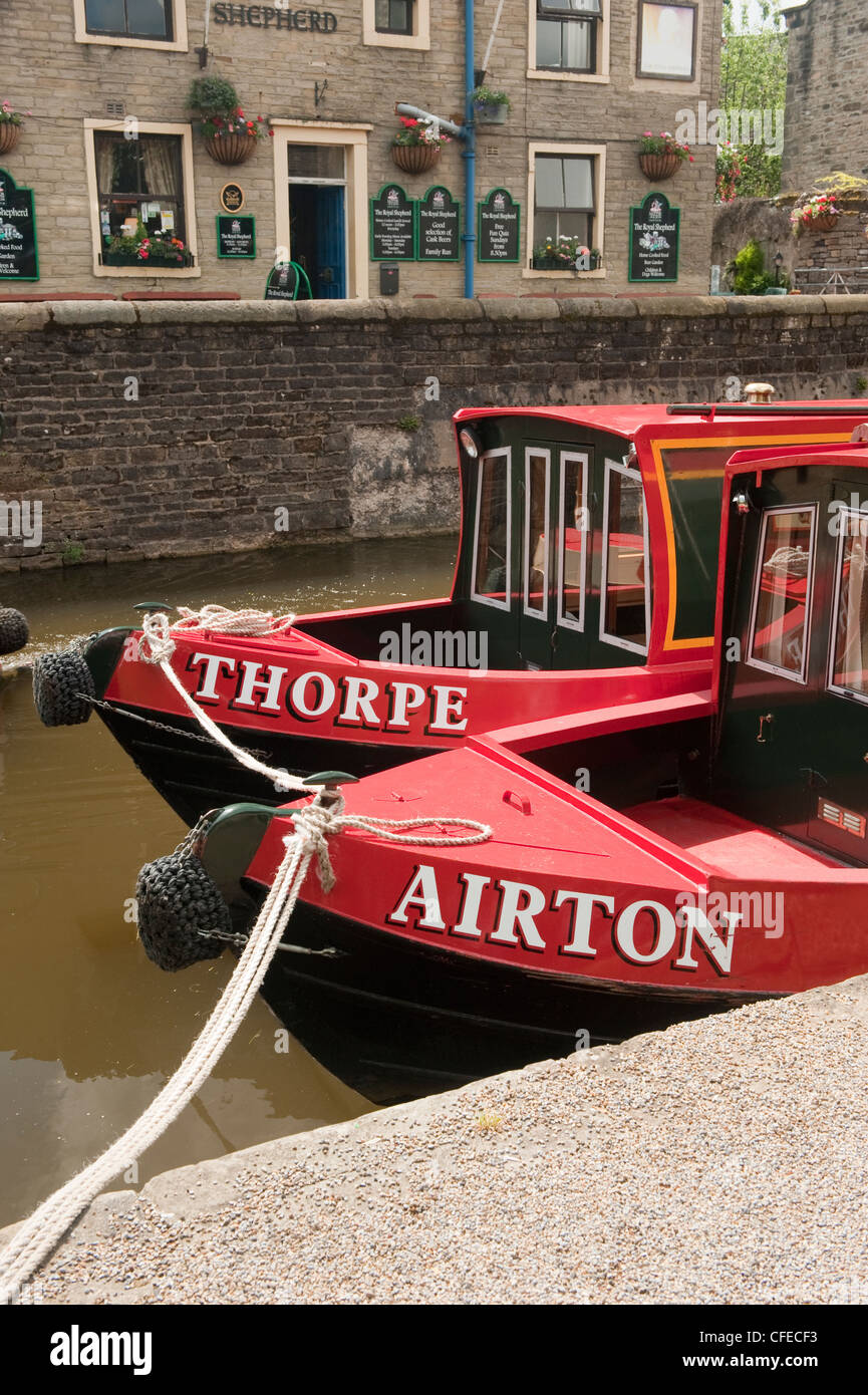 Face à 2 bateaux de canal rouge vif (noms sur l'arc) amarré côte à côte par le chemin de remorquage et le pub canalside - Canal Leeds-Liverpool, Skipton, Yorkshire, Angleterre. Banque D'Images