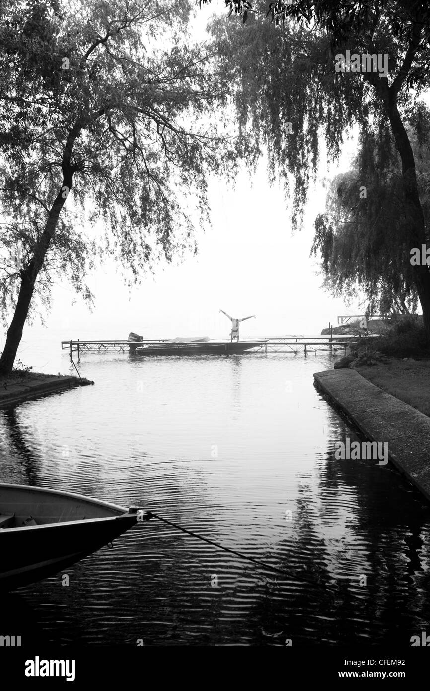 Silhouette en noir et blanc d'une fille cartwheeling sur un petit bateau, quai, arbres et de l'eau ridée encadrent son chiffre. Banque D'Images