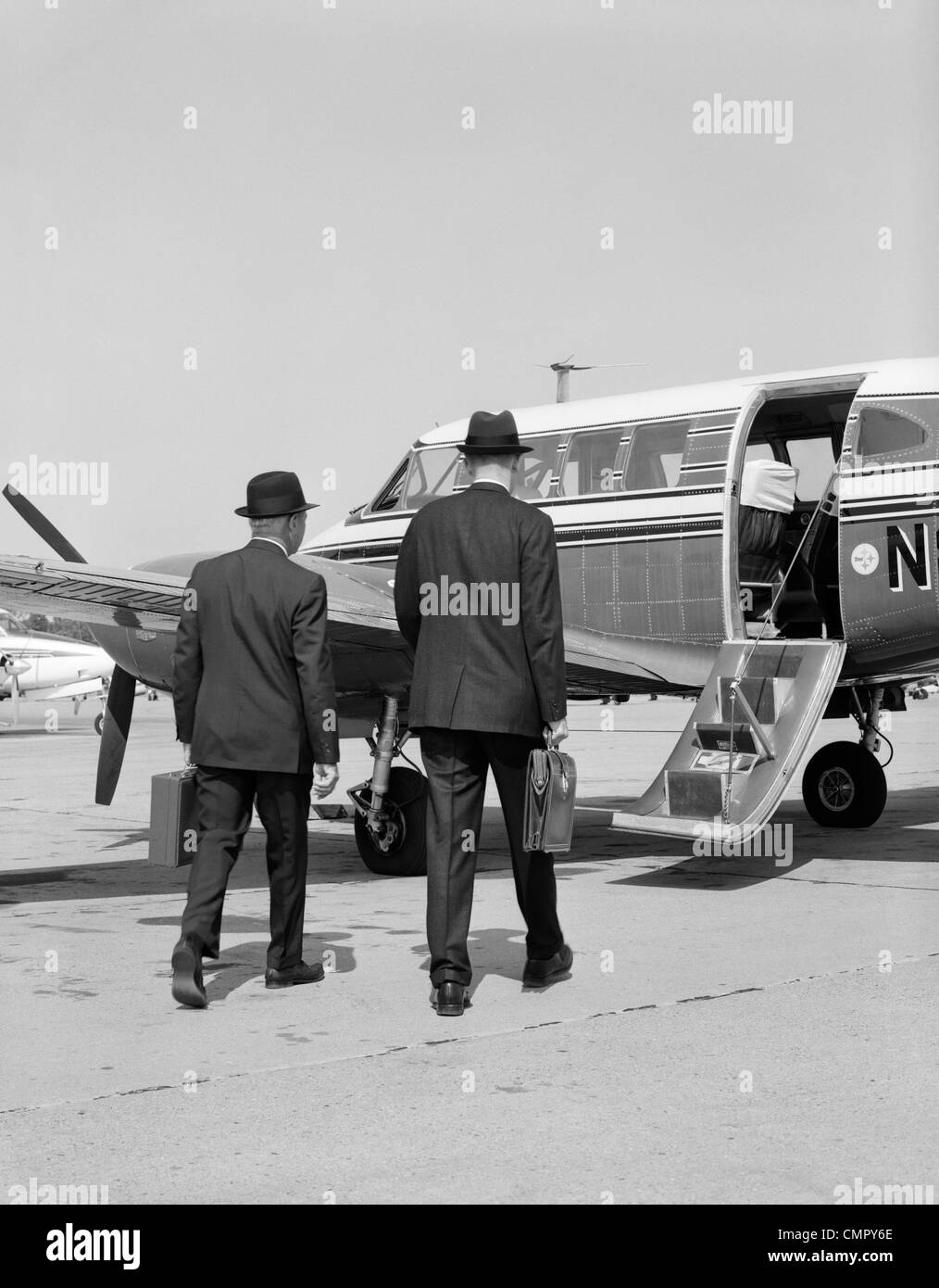 1960 DEUX HOMMES D'AFFAIRES À MARCHER ENSEMBLE SUR UNE PETITE MALLETTE DE TRANSPORT AVION hélice Banque D'Images