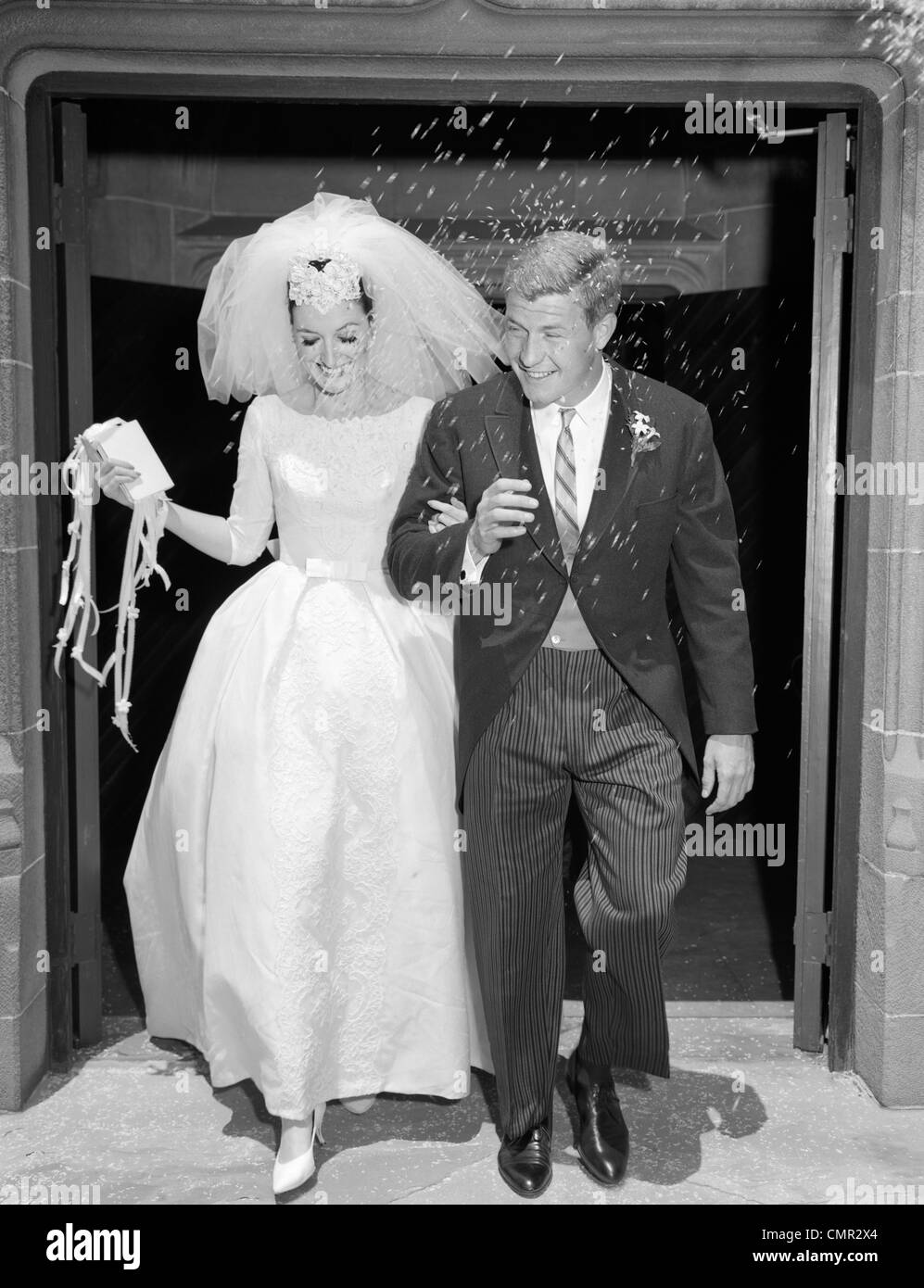 1960 COUPLE MARIÉS DANS LES BRAS LAISSANT ÉGLISE SOUS DOUCHE DE RIZ Banque D'Images