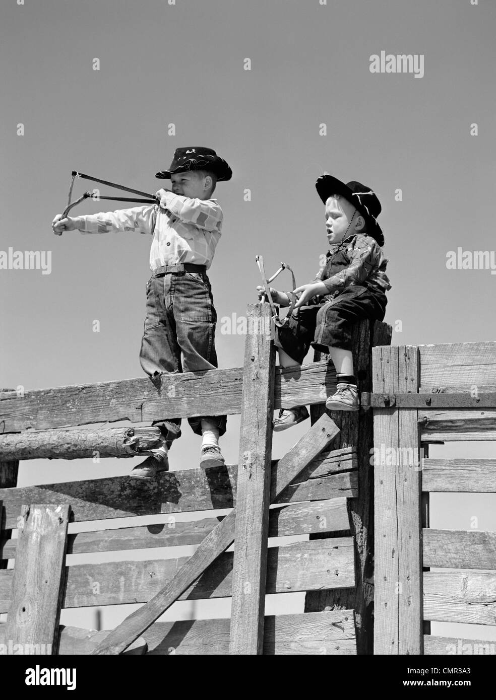 Années 50, deux jeunes garçons habillés comme des cowboys de frondes de TIR AU SOMMET D'une clôture de bois Banque D'Images