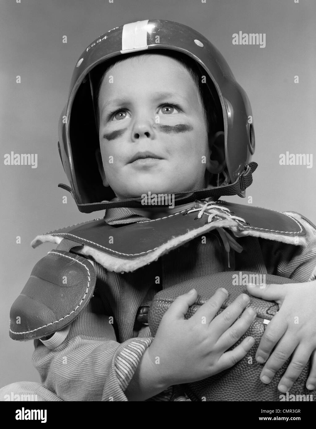 1950 PORTRAIT DE GARÇON EN CASQUE ET ÉPAULETTES PORTANT LA GRAISSE DE PEINTURE SOUS LES YEUX & HOLDING FOOTBALL Banque D'Images