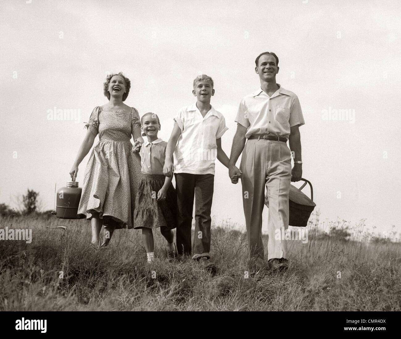 1950 FAMILY WALKING IN GRASSY MEADOW exerçant son panier de pique-nique de l'été et le Thermos Banque D'Images