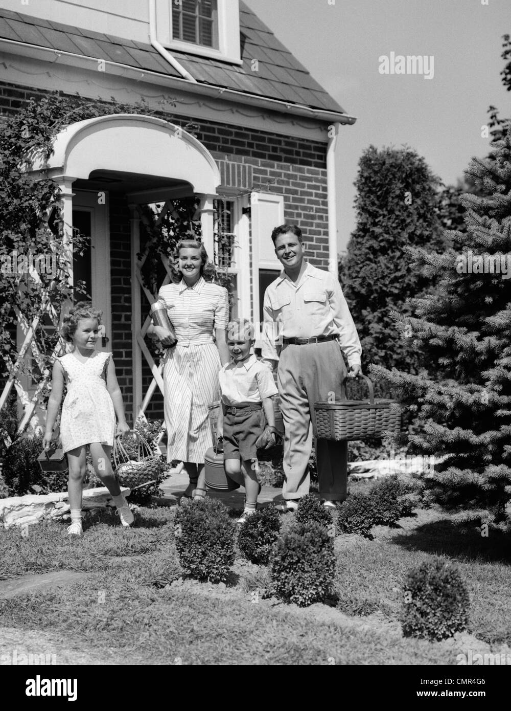 Années 1940 Années 1950 FAMILLE DE QUATRE MARCHER HORS DE LA MAISON PORTANT DES PANIERS DE PIQUE-NIQUE ET THERMOS VERSEUSE Banque D'Images