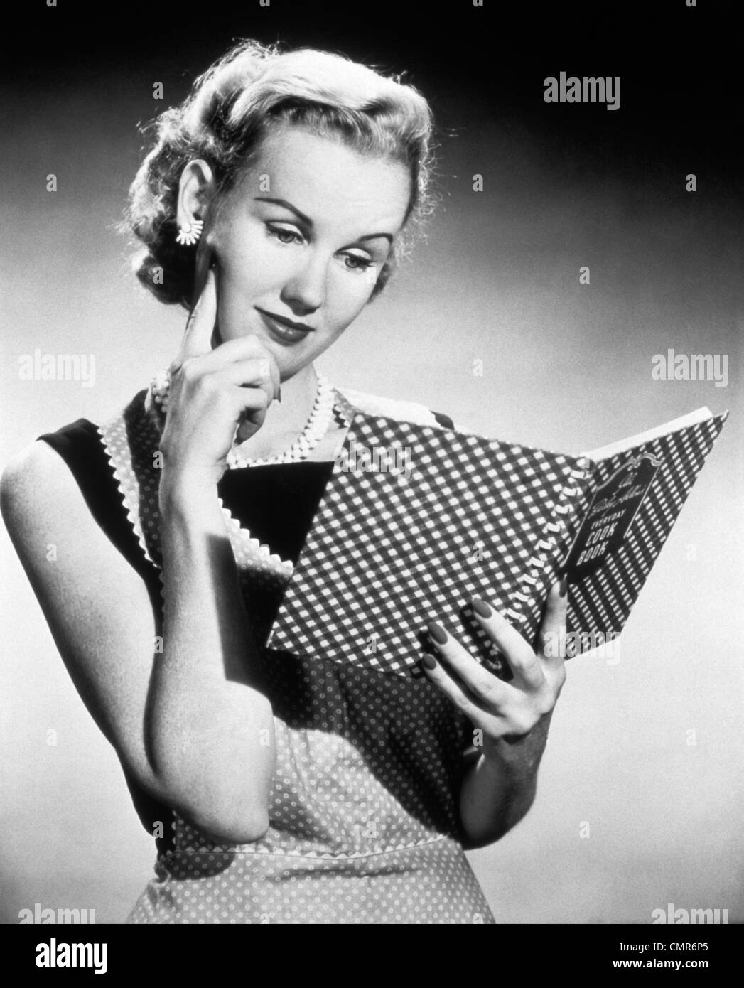 1950 FEMME AU FOYER BLONDE RÉFLÉCHIE LA LECTURE DE LIVRE de cuisinier Banque D'Images