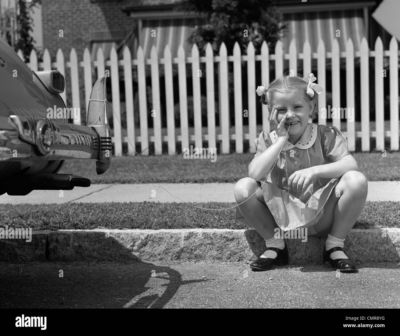 1940 GIRL SITTING ON CURB AVEC TOOTH LIÉE AU RETOUR D'AILE DE VOITURE LOOKING AT CAMERA Banque D'Images