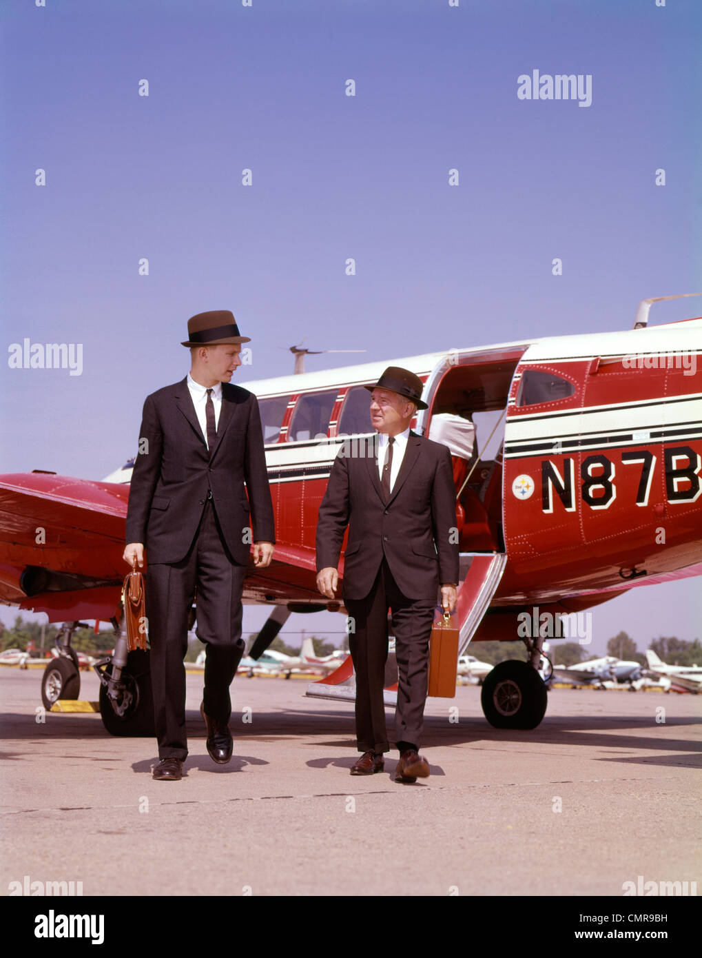 1960 DEUX HOMMES EN COSTUMES ET CHAPEAUX EXERÇANT SON PORTE-DOCUMENTS À PIED DE UNE PETITE CHAMBRE LITS JUMEAUX D'AFFAIRES PARLER AVION PROP Banque D'Images