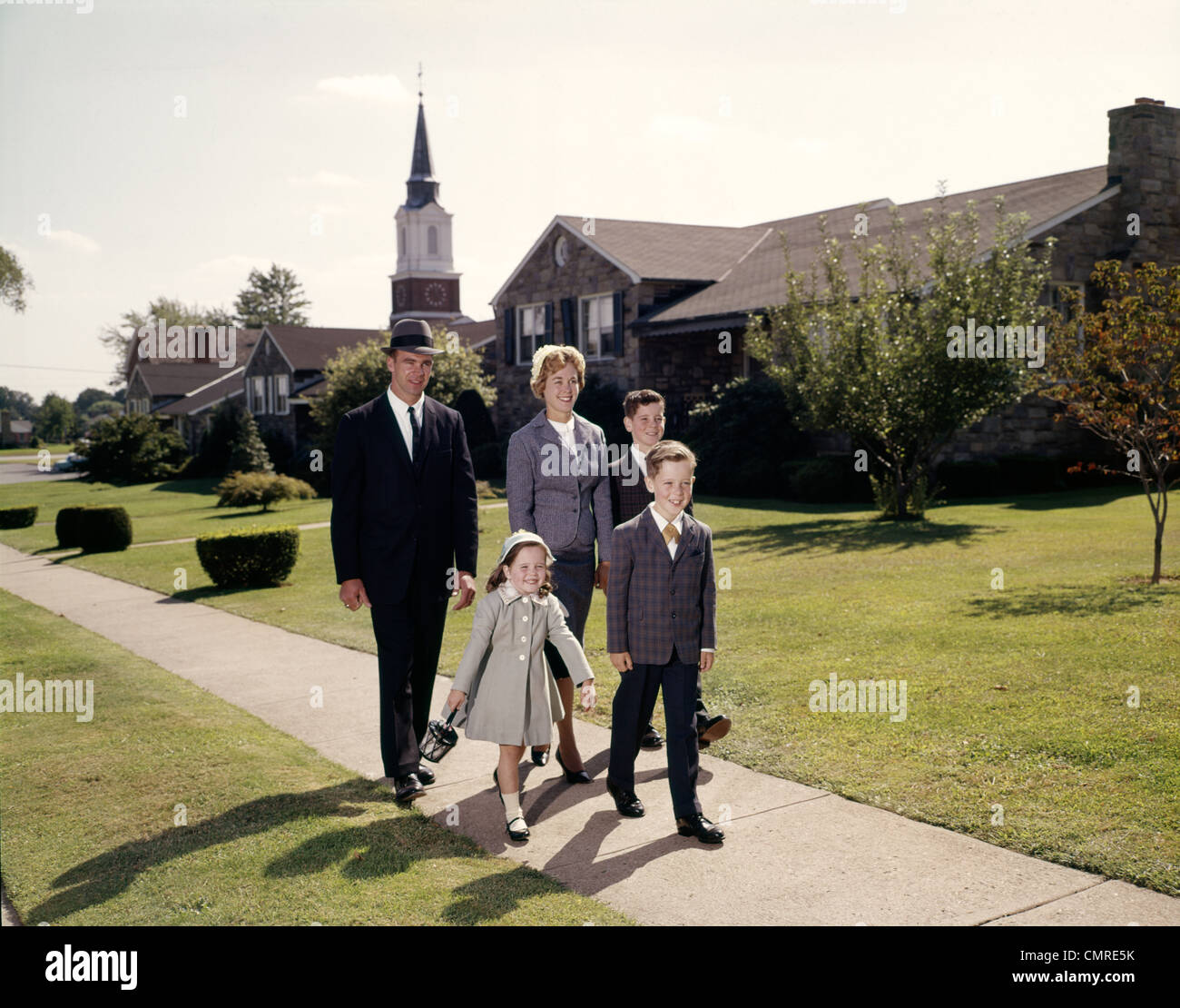 1960 FAMILLE DE 5 MARCHE SUR UN TROTTOIR DE BANLIEUE CLOCHER DE L'ÉGLISE EN ARRIÈRE-PLAN Banque D'Images