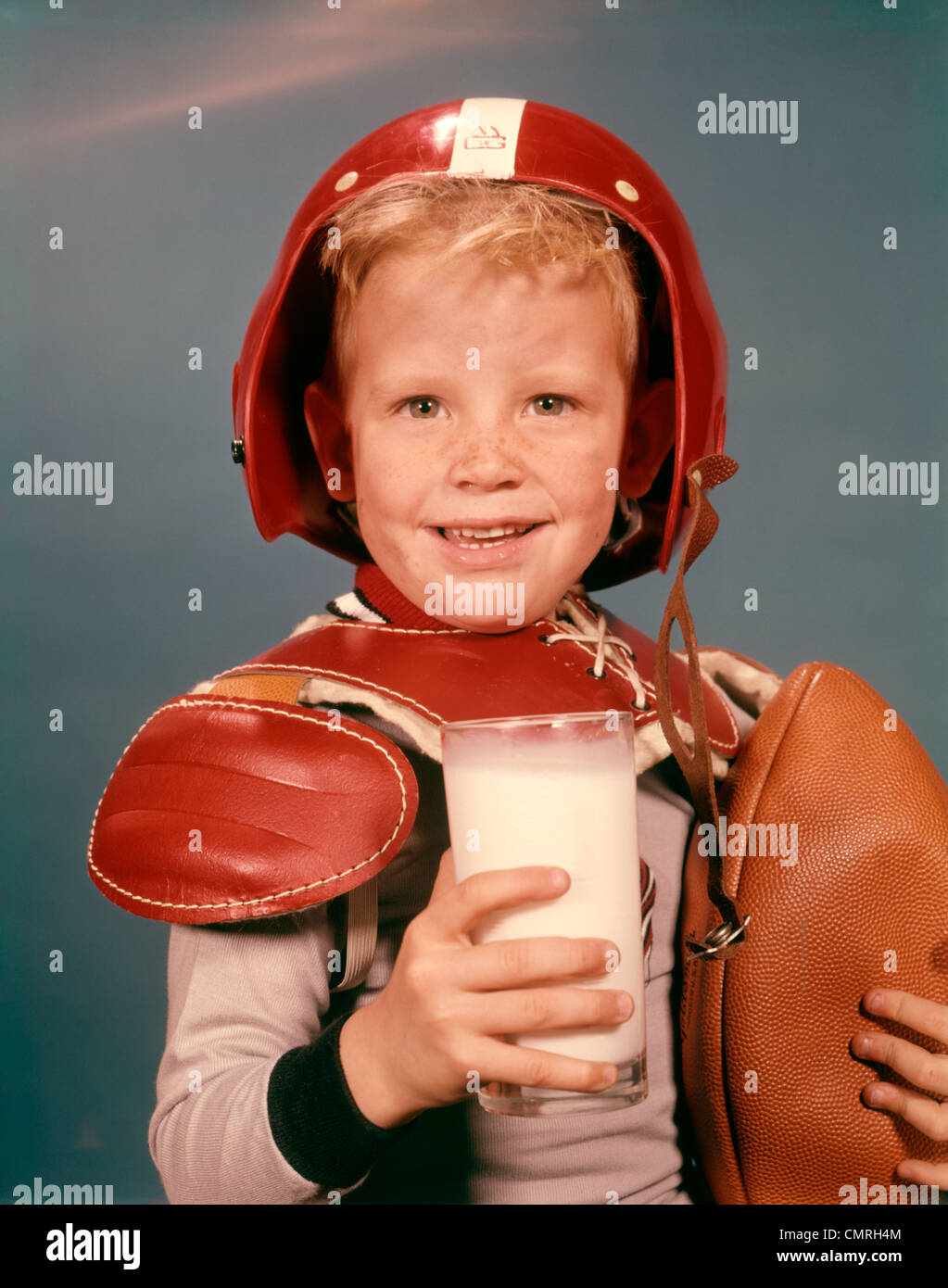 1960 BOY SMILING BLONDE WEARING RED HELMET FOOTBALL SHOULDER PADS HOLDING GLASS LE LAIT ET LE FOOTBALL LOOKING AT CAMERA Banque D'Images