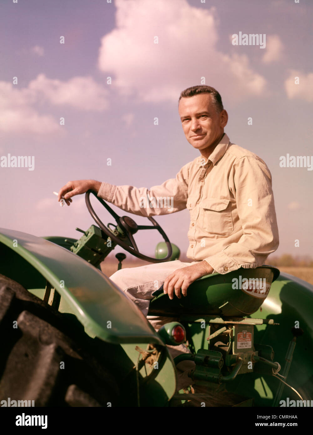 1960 PORTRAIT SMILING FARMER SITTING ON CIGARETTE TRACTEUR VERT Banque D'Images