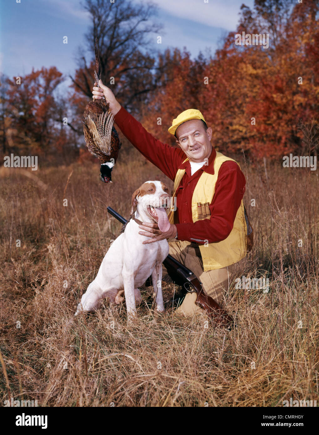 Années 1950 Années 1960 MAN HUNTER AVEC FUSIL HOLDING UP BIRD PHEASANT AGENOUILLÉE À CÔTÉ DE CHIEN DE CHASSE À L'AUTOMNE Banque D'Images