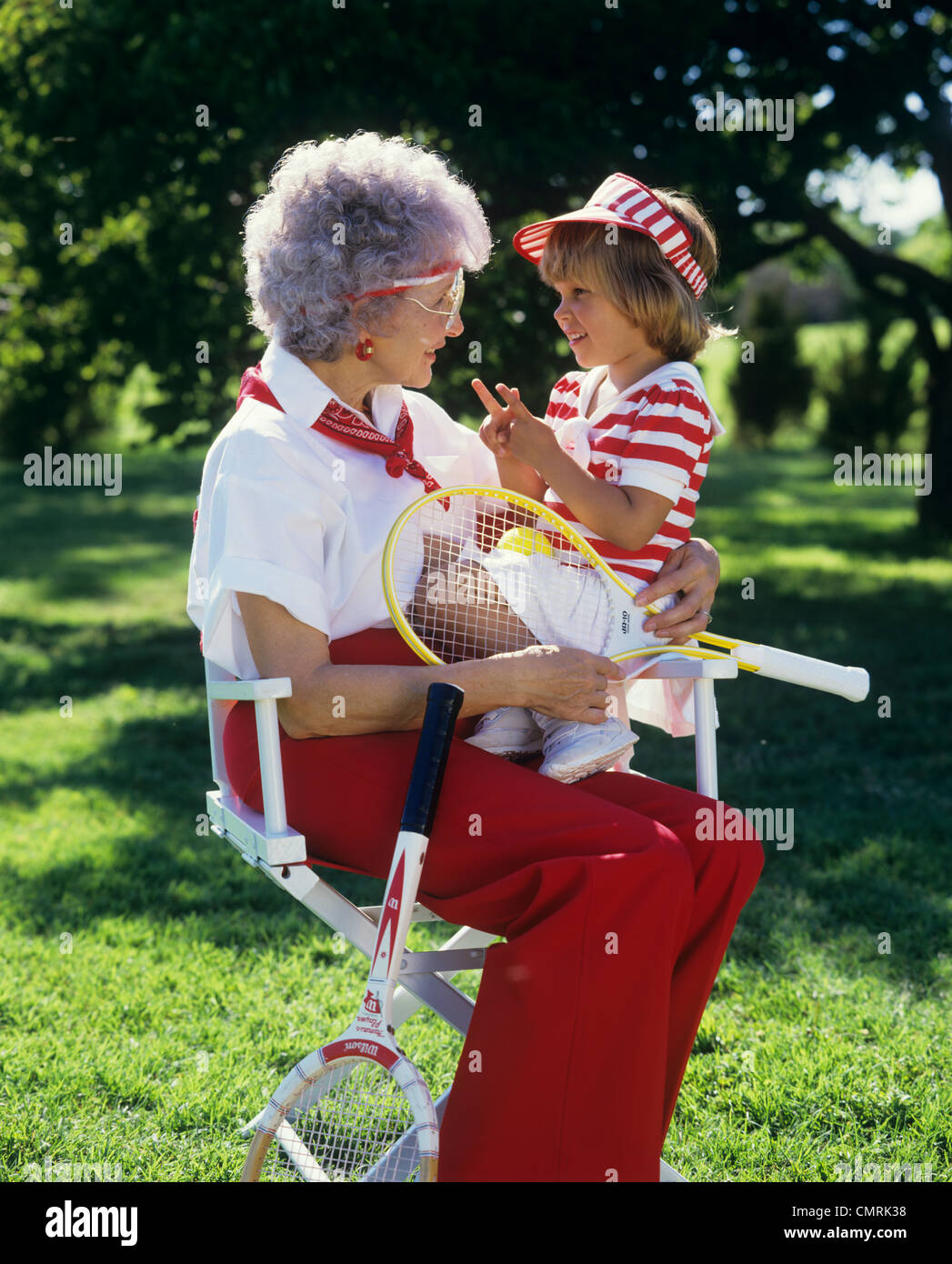 1980 GRAND-MÈRE ASSISE DANS UNE CHAISE HOLDING raquette de tennis et sa petite-fille portant des vêtements de tennis Banque D'Images