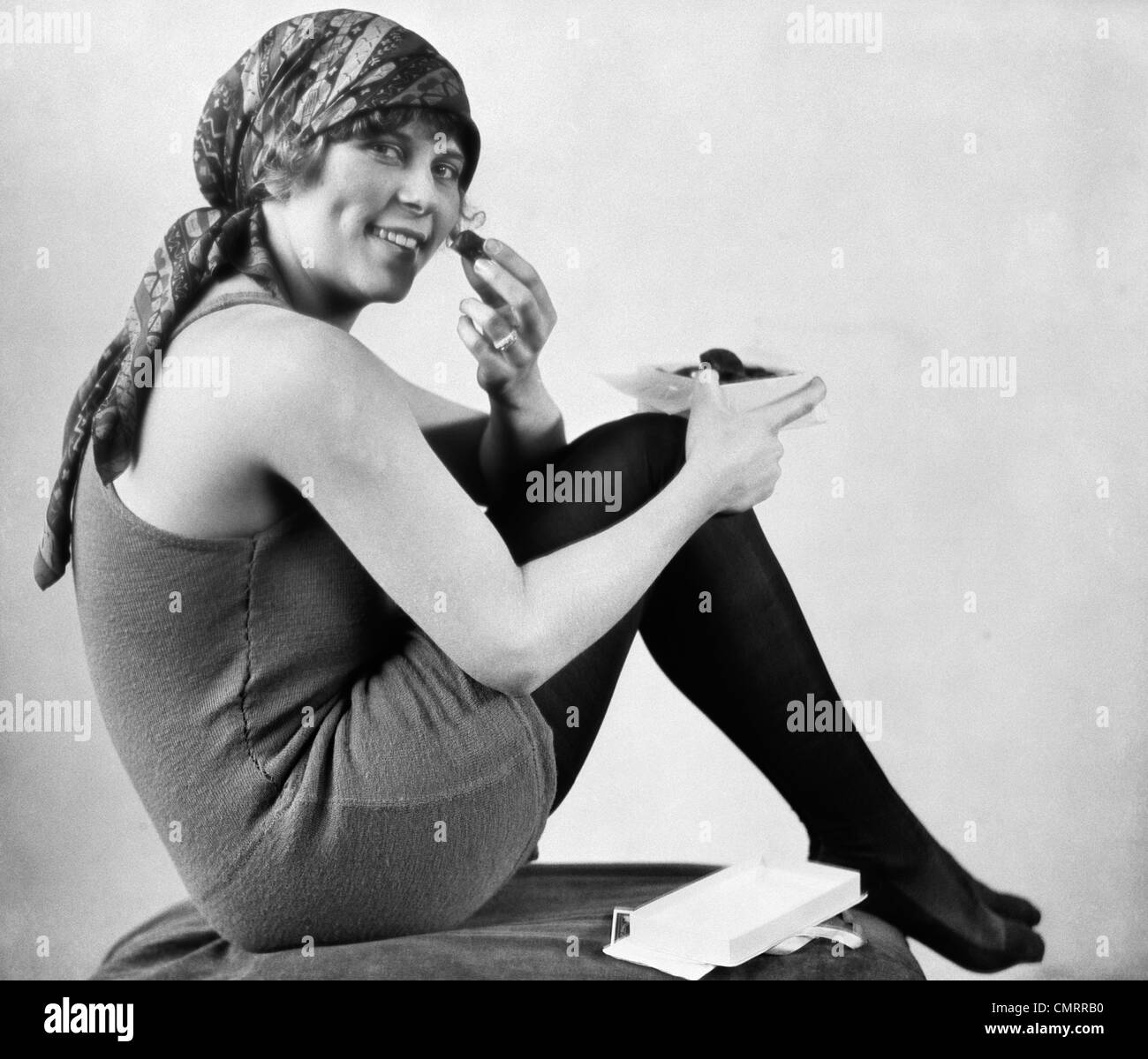 Années 1920 femme en maillot de bain à l'ANCIENNE AVEC FOULARD SUR LA TÊTE ASSIS AVEC LES GENOUX À LA POITRINE DE MANGER DES CHOCOLATS LOOKING AT CAMERA Banque D'Images
