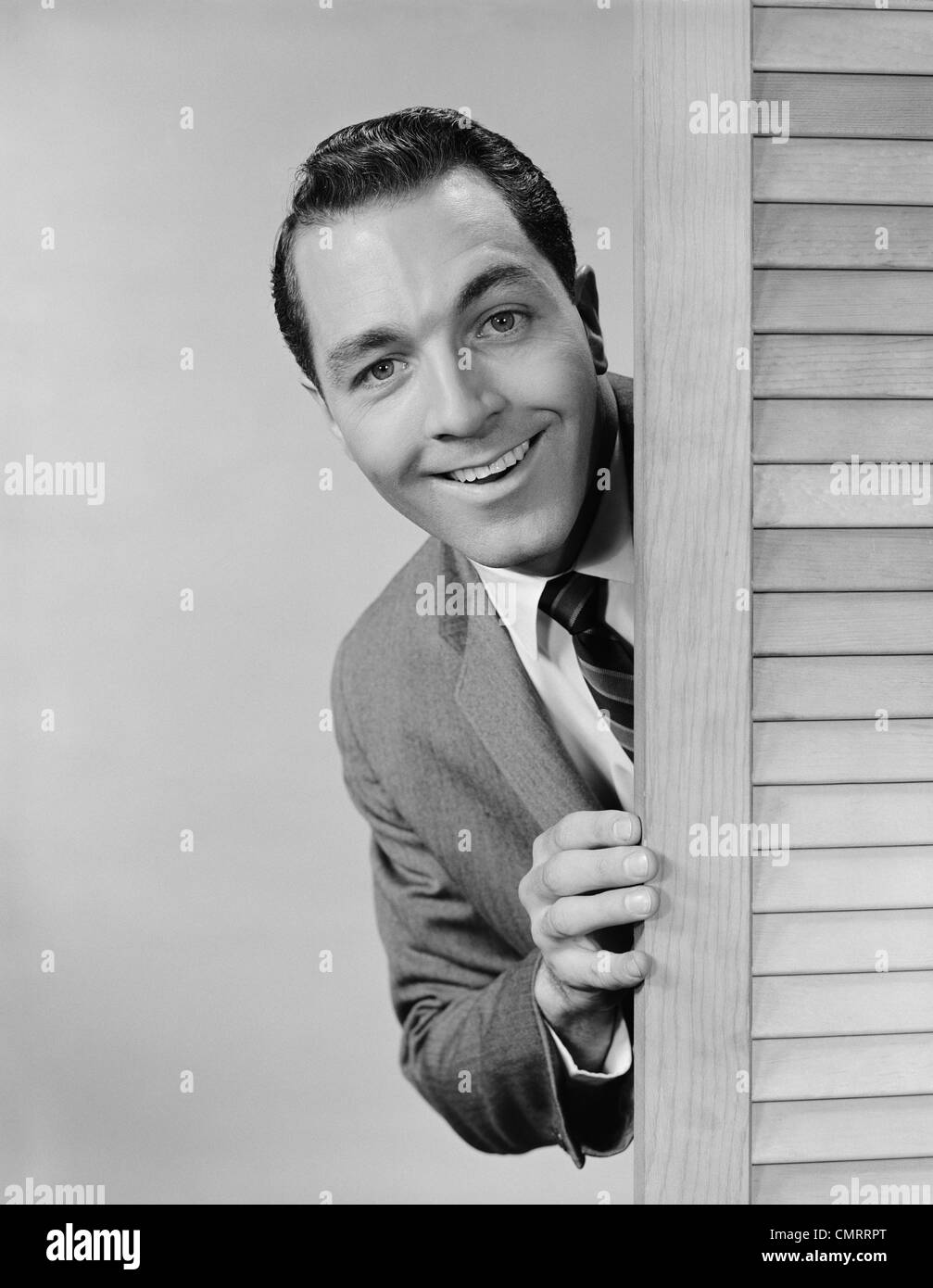 1960 SMILING MAN PEEKING COSTUME D'AFFAIRES DE DERRIÈRE LA PORTE À PERSIENNES Banque D'Images