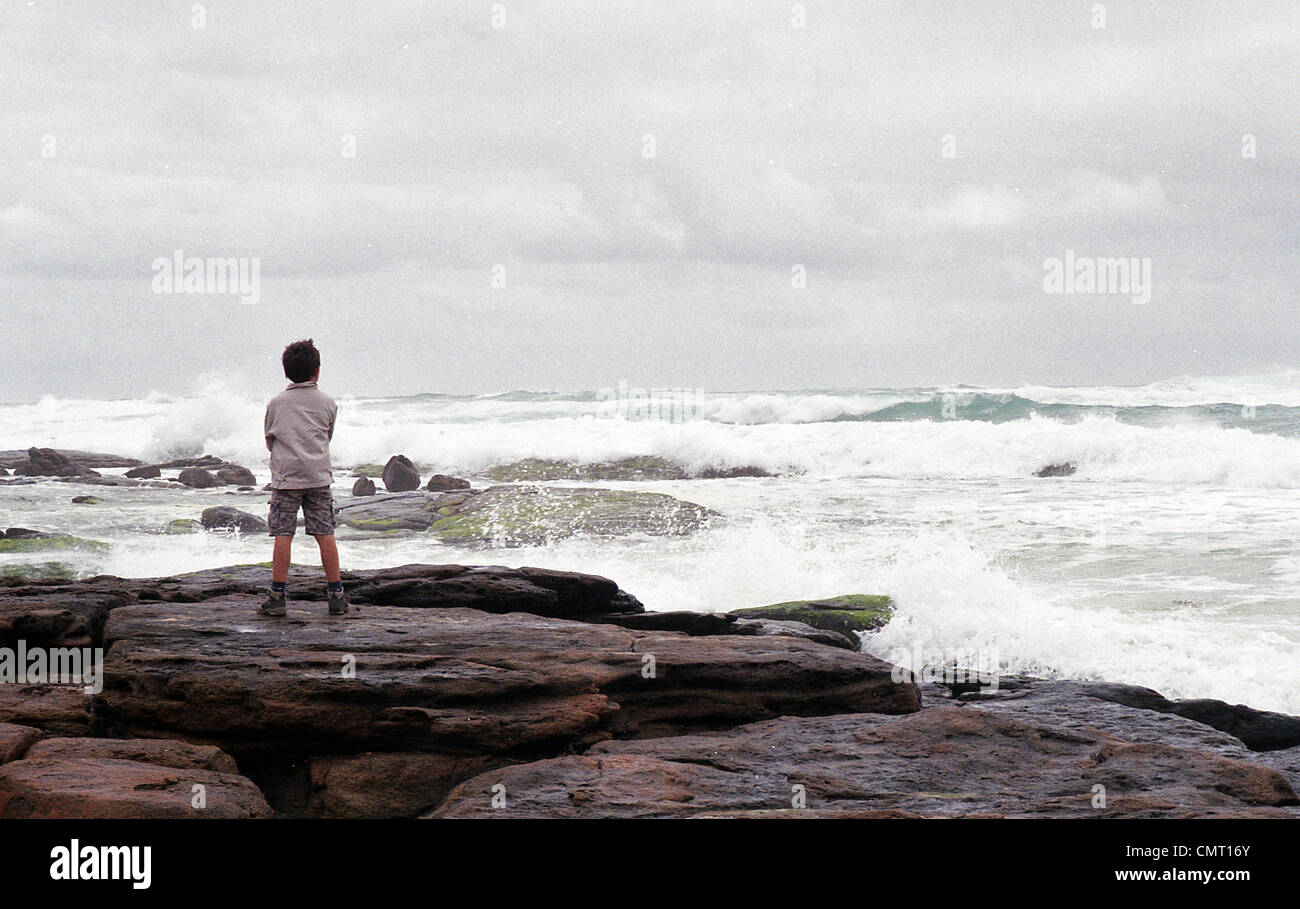 Un garçon se dresse sur des rochers face à une mer déchaînée et d'appréhension des nuages Banque D'Images
