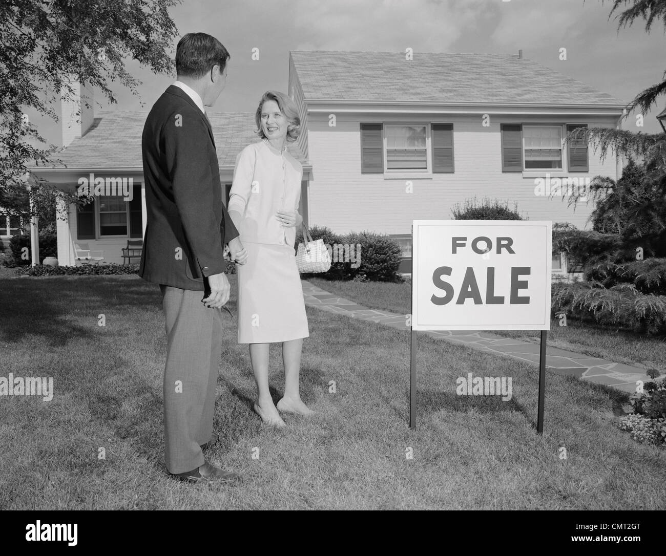 1960 HOMME FEMME COUPLE standing in front of house avec FOR SALE SIGN IN YARD Banque D'Images