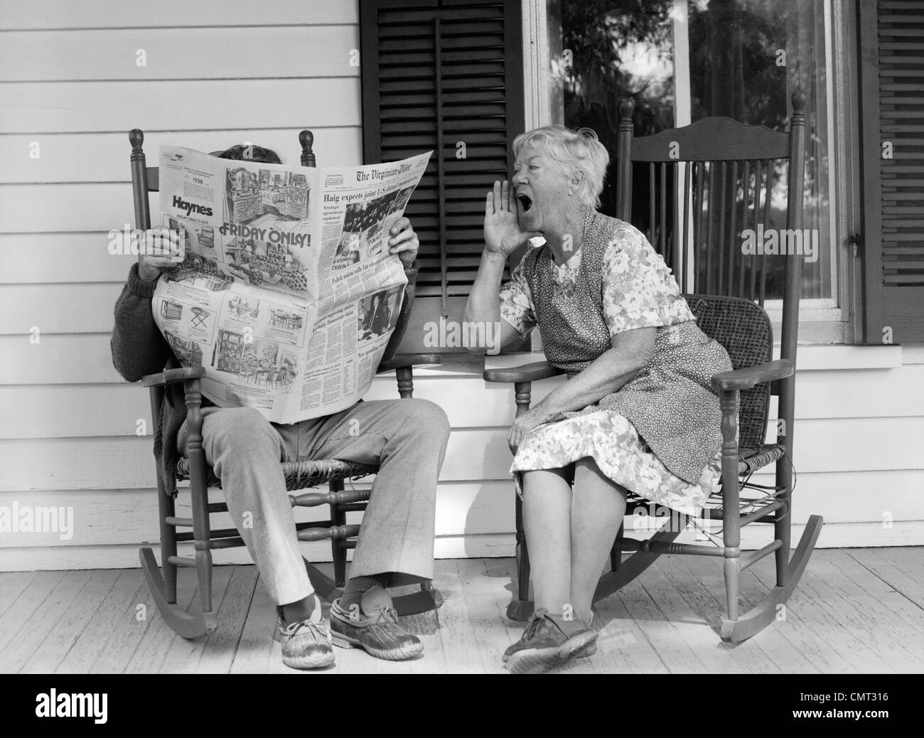 1970 COUPLE DE PERSONNES ÂGÉES DANS DES CHAISES À BASCULE SUR LE PORCHE MAN READING NEWSPAPER TANDIS QU'épouse se lamente sur lui Banque D'Images
