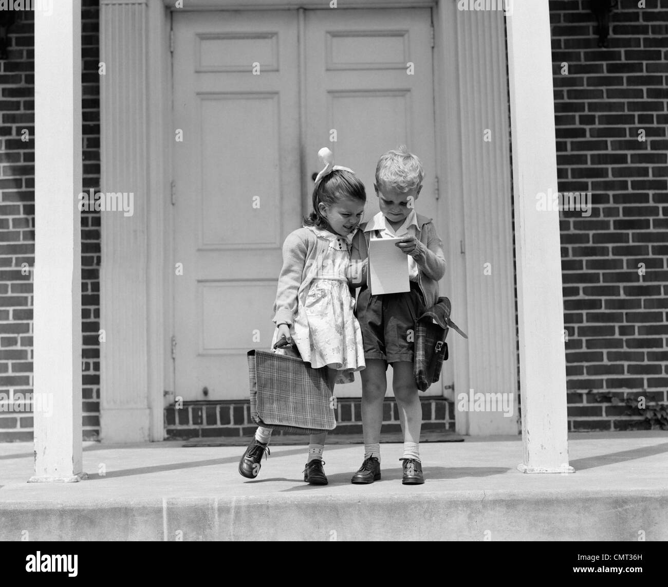 1940 petit garçon et fille avec sacs d'ÉCOLE À LA RECHERCHE À L'EXTÉRIEUR DU BLOC-NOTES Banque D'Images