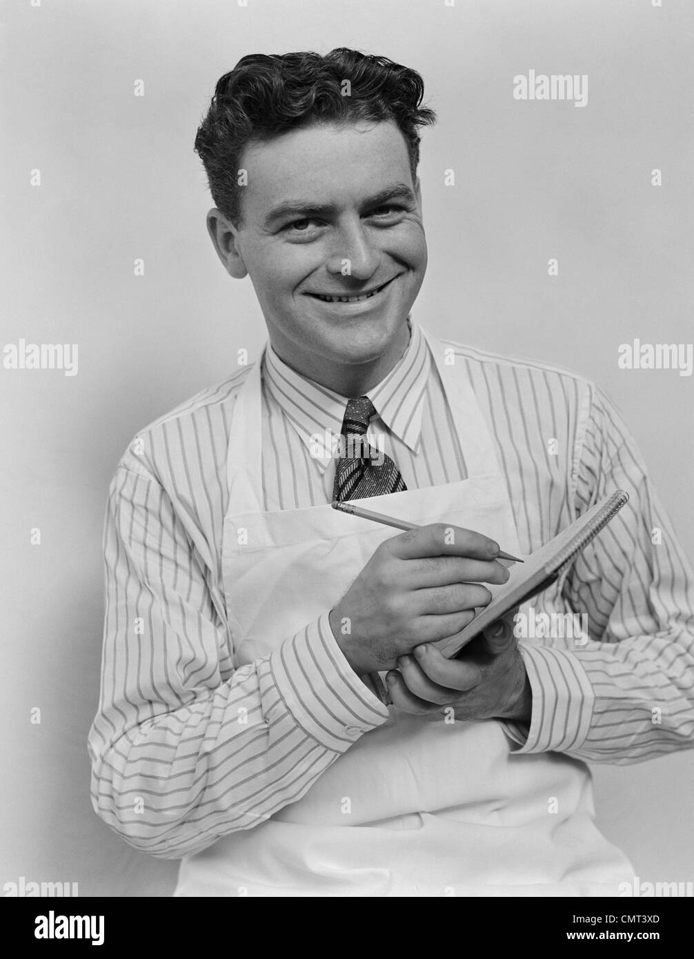 Années 1930 Années 1940 cheveux bouclés SMILING MAN WEARING APRON TAKING ORDER AVEC CRAYON ET NOTES LOOKING AT CAMERA Banque D'Images