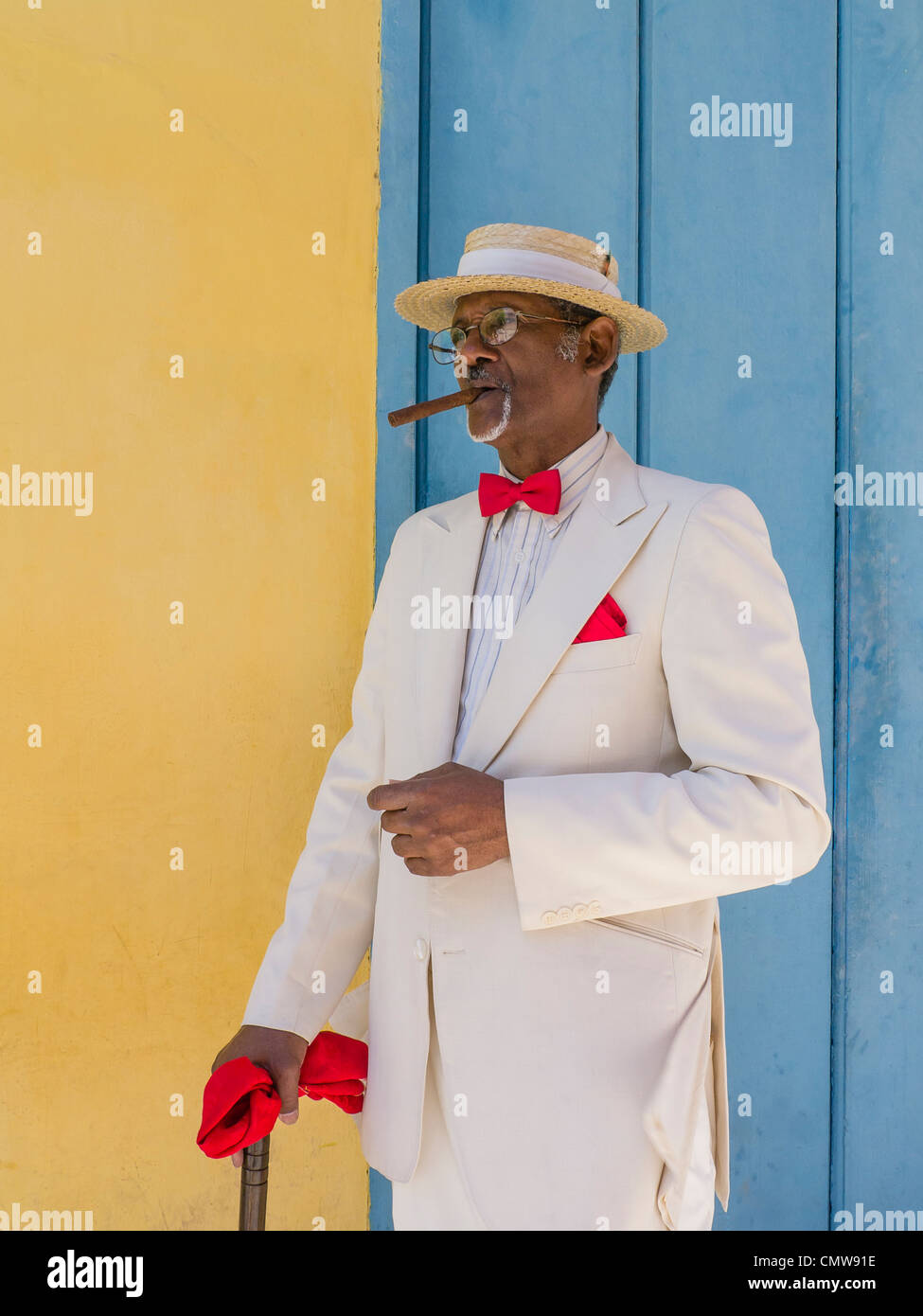 Un représentant de Cuba Personnes âgées sombre dans un canotier de paille  chapeau, costume blanc, rouge noeud papillon, et un cigare se distingue par  un mur coloré à La Havane Photo Stock -