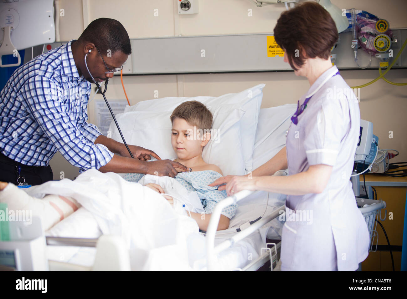 Un jeune garçon est contrôlé par un médecin et l'infirmière sur un hôpital pour enfants ward après une opération UK Banque D'Images