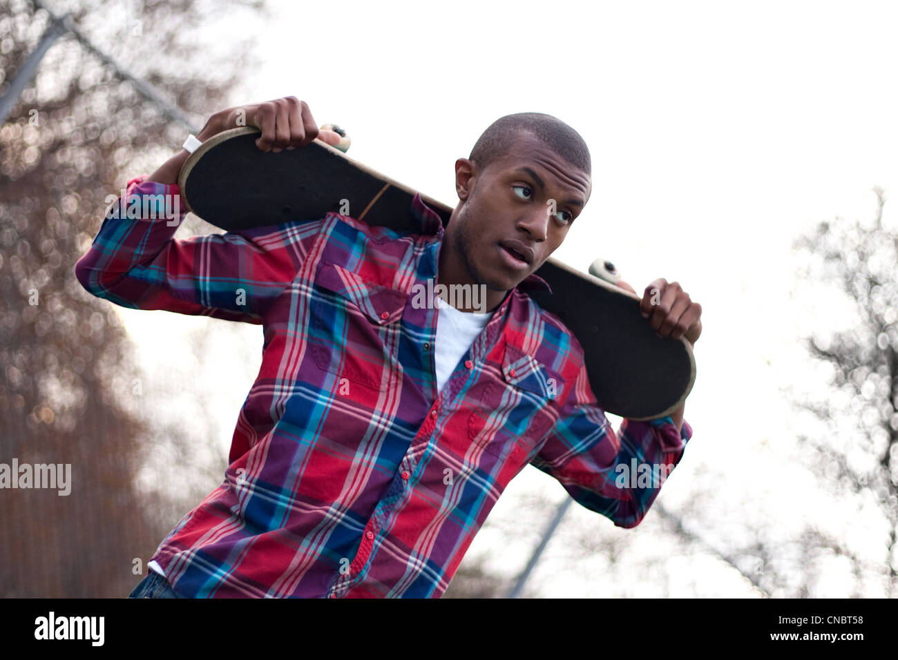 Un jeune homme traînant en tenant son skateboard sur l'arrière de ses épaules au-dessus de sa tête. Banque D'Images
