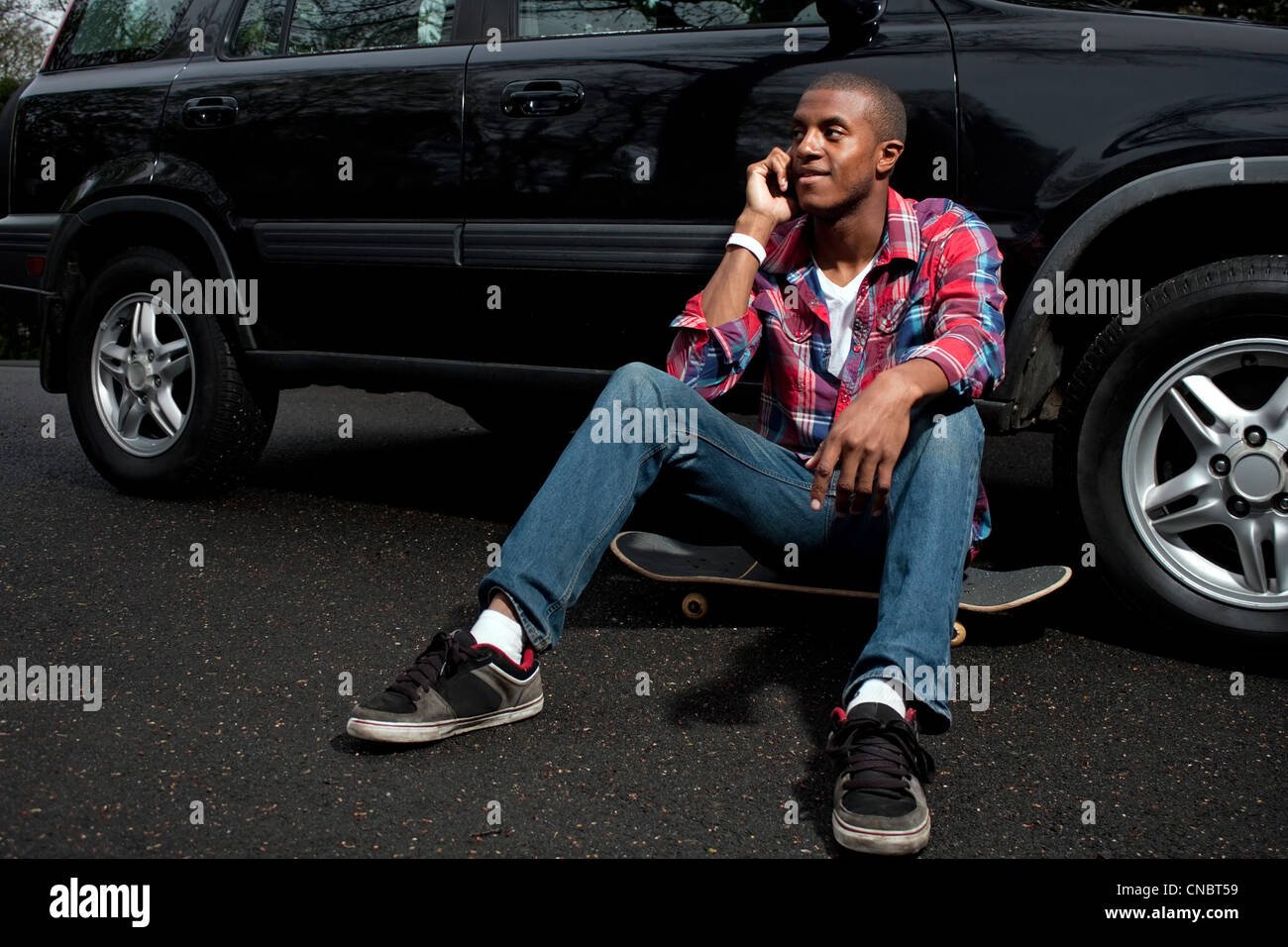 Un jeune homme traînant assis sur son skateboard près de son véhicule comme il parle sur son smartphone. Banque D'Images