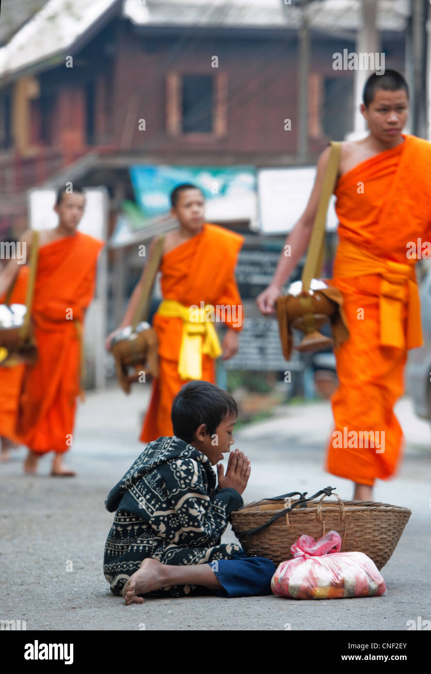 Jeune garçon Lao pour quémander l'aumône de passer les moines bouddhistes qui viennent de recevoir l'aumône pendant matin procession d'aumône Banque D'Images