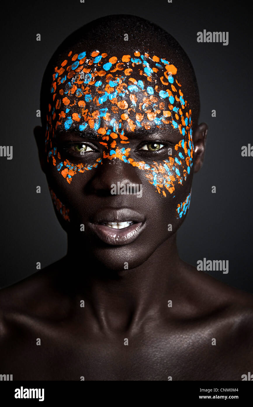 Les jeunes Noirs africains modèle masculin vêtu d'orange et de bleu créatif peinture sur visage tribal, photographiés dans un environnement de studio. Banque D'Images