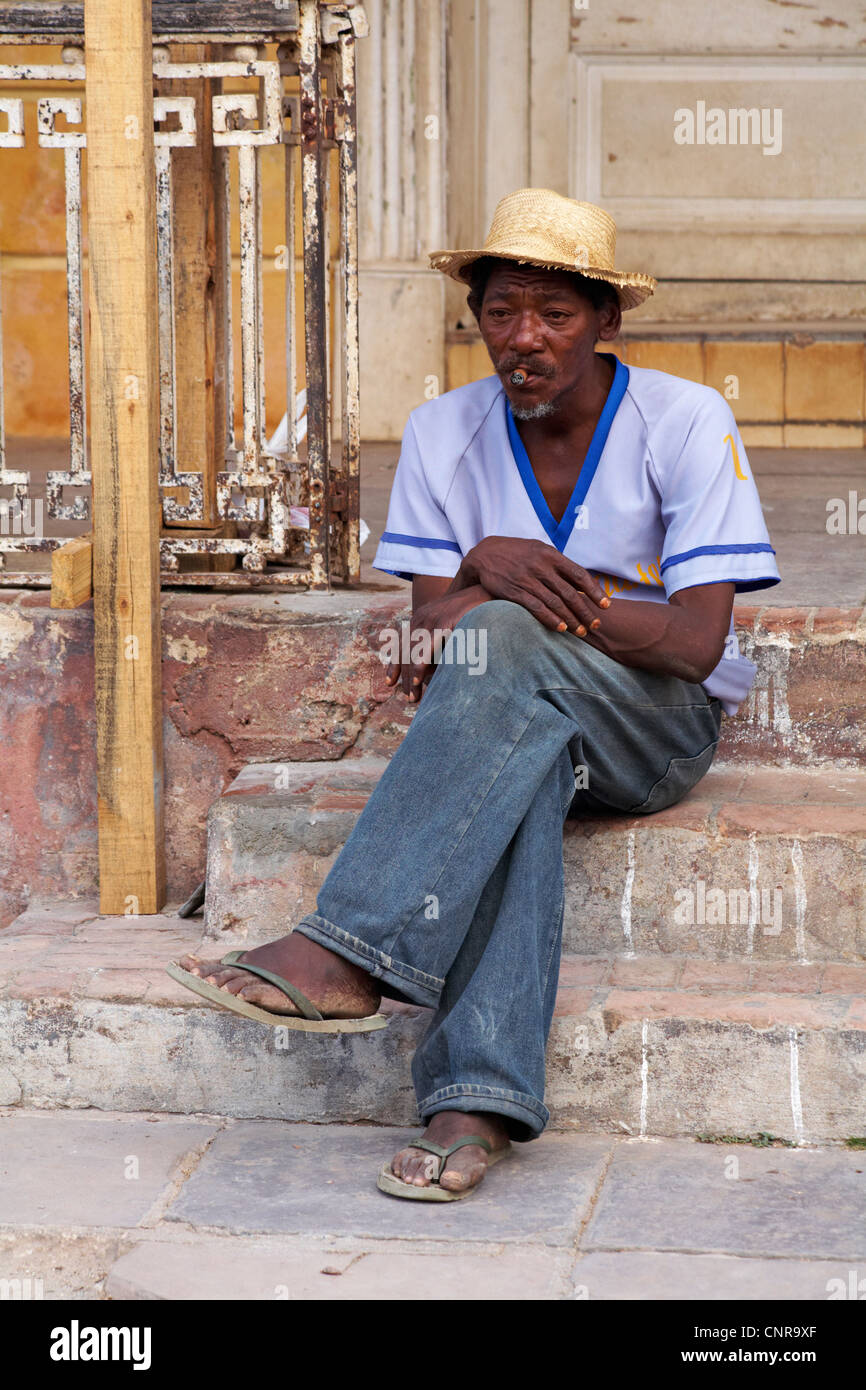 La vie quotidienne à Cuba - afro-caraïbes homme assis sur les marches fumer un cigare à Trinidad, Cuba Banque D'Images