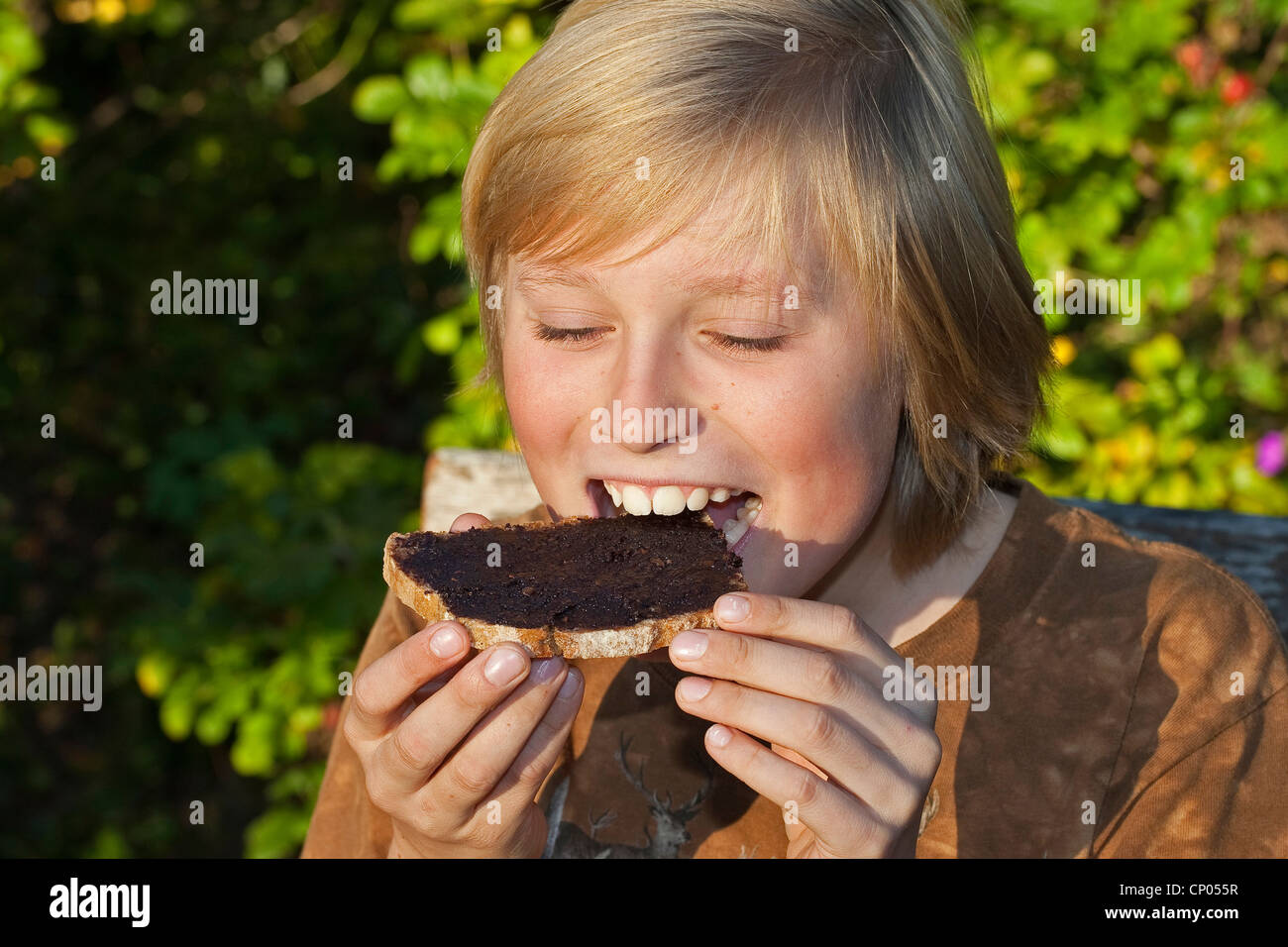 Le noisetier commun (Corylus avellana), garçon assis à la table de jardin de manger une tranche de pain avec pâte à tartiner au chocolat qu'il a faite de l'auto-prélèvement de noisettes, la poudre de cacao, le beurre et le sucre, Allemagne Banque D'Images