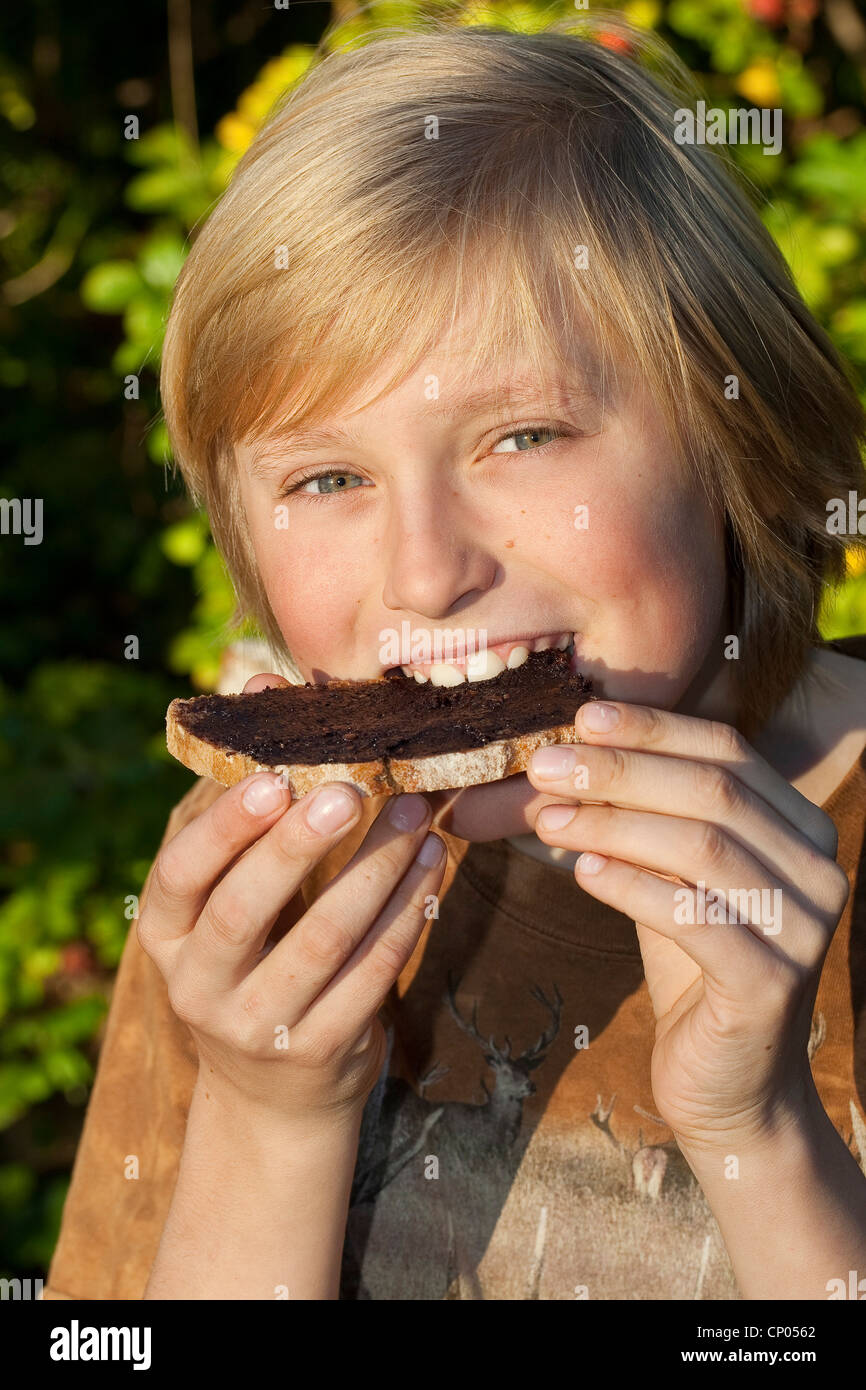 Le noisetier commun (Corylus avellana), garçon assis à la table de jardin de manger une tranche de pain avec pâte à tartiner au chocolat qu'il a faite de l'auto-prélèvement de noisettes, la poudre de cacao, le beurre et le sucre, Allemagne Banque D'Images