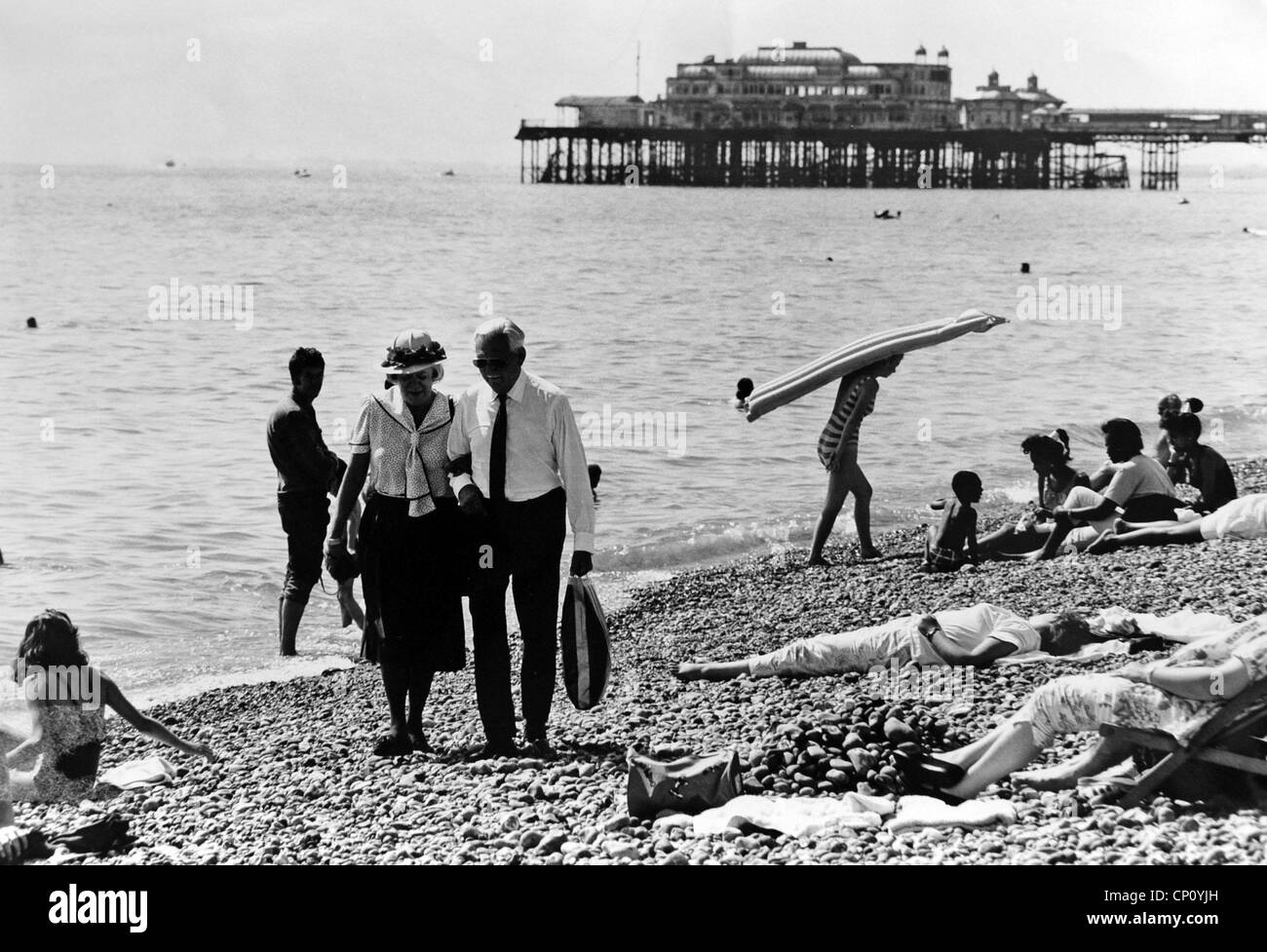 Couple smartly dressed marcher le long de la plage de Brighton parmi les baigneurs avec le West Pier en arrière-plan vers 1983 Banque D'Images