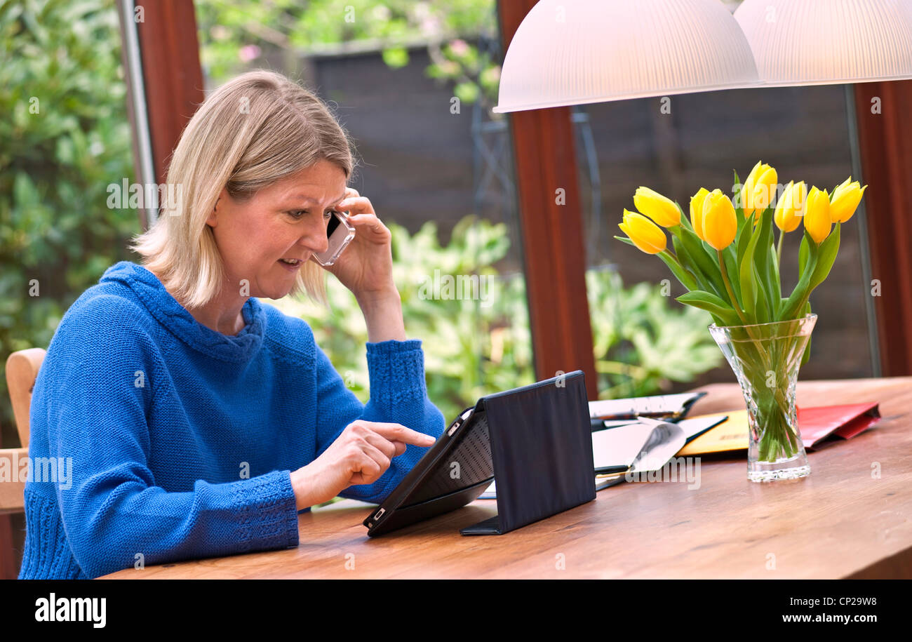 Femme blonde qui travaille à la maison dans le jardin d'hiver à l'aide de son iPad tablet computer et parler sur son smartphone iPhone Banque D'Images