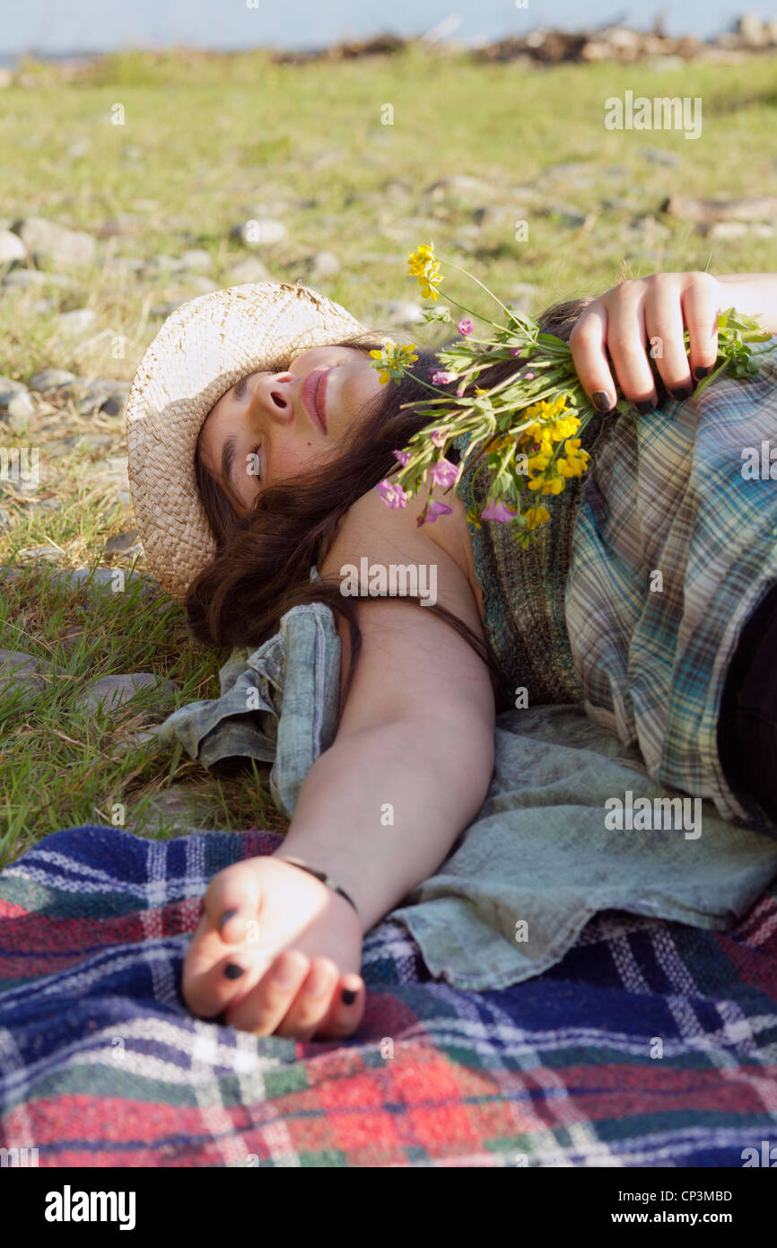 Jeune fille avec chapeau de soleil se trouve dormant sur une couverture, tenant des fleurs dans sa main. L'été ensoleillé tourné en extérieur. Banque D'Images