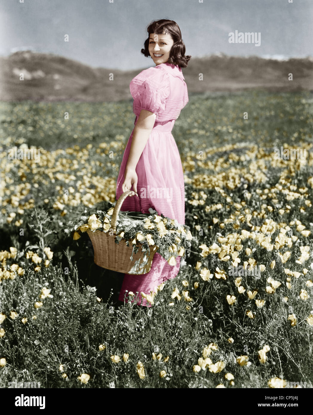 Vue arrière Girl standing in meadow holding panier de fleurs smiling Banque D'Images