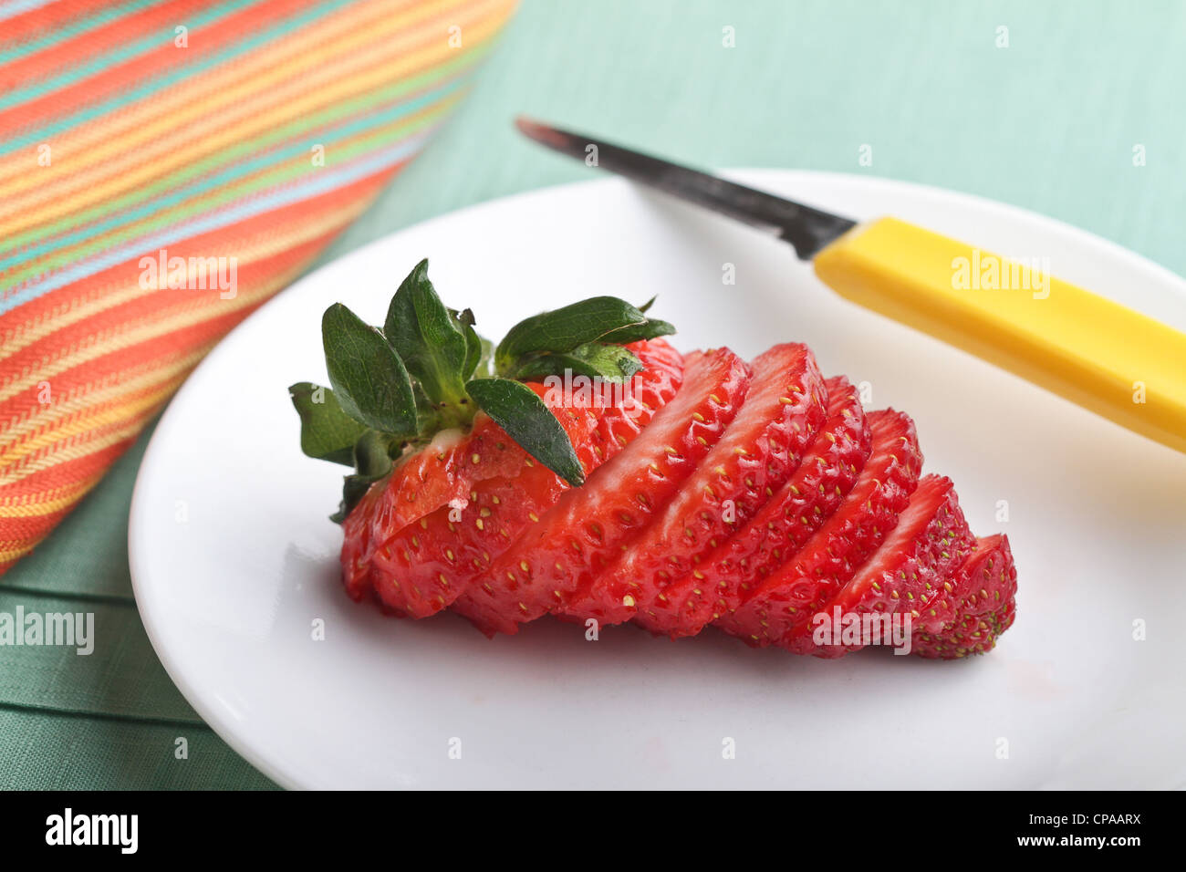 Un ensemble de fraises fraîches, tranchées sur une plaque blanche sur un endroit vert mat avec une serviette à rayures jaune et couteau à légumes. Banque D'Images