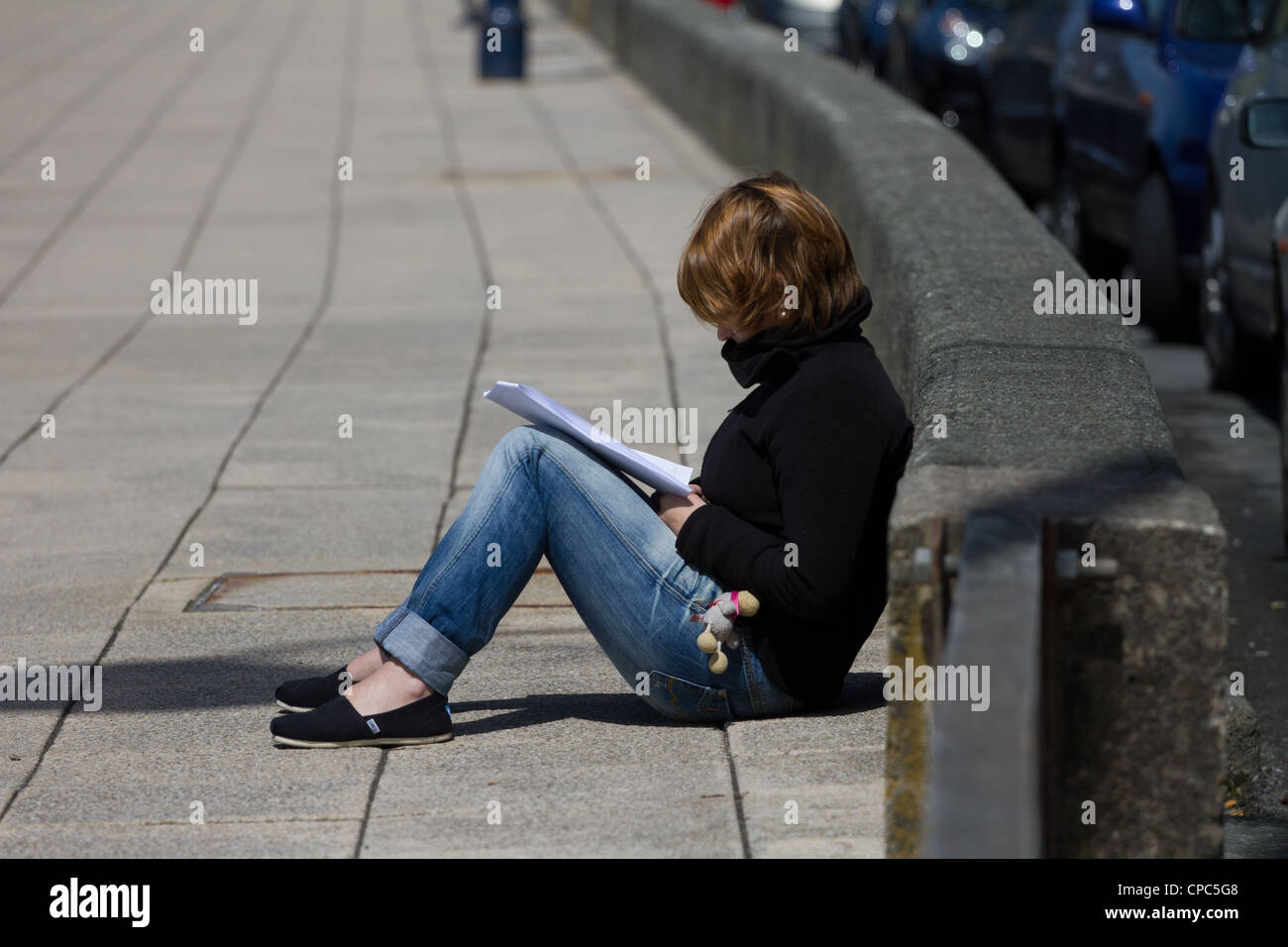 Une jeune étudiante de la révision au soleil assis sur un trottoir et appuyé contre un mur bas. Banque D'Images