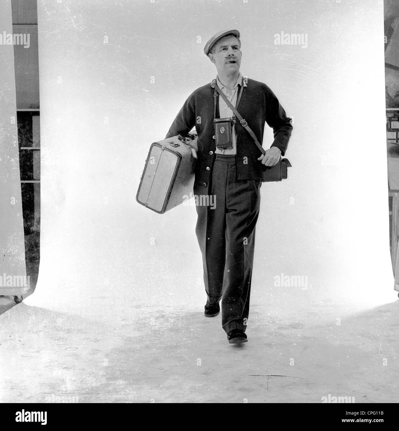 Publicité 1960s. Homme dans un studio photograhique debout contre un écran blanc agissant comme un holographe avec sa valise, appareil photo et sac à bandoulière. Banque D'Images