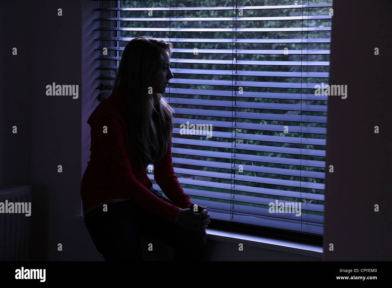Jeune femme assise seule dans une pièce sombre à la recherche à travers une fenêtre aveugle. Banque D'Images