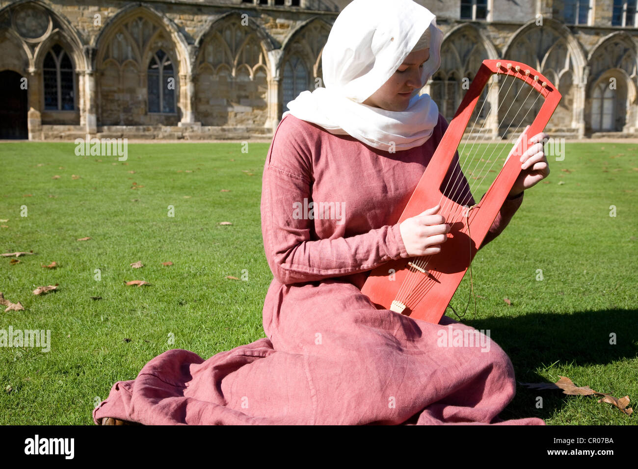 UK. L'Angleterre. Battle Abbey. East Sussex. Femme en costume médiéval à l'extérieur assis sur l'herbe pelouse. La lecture de musique traditionnelle inst Banque D'Images