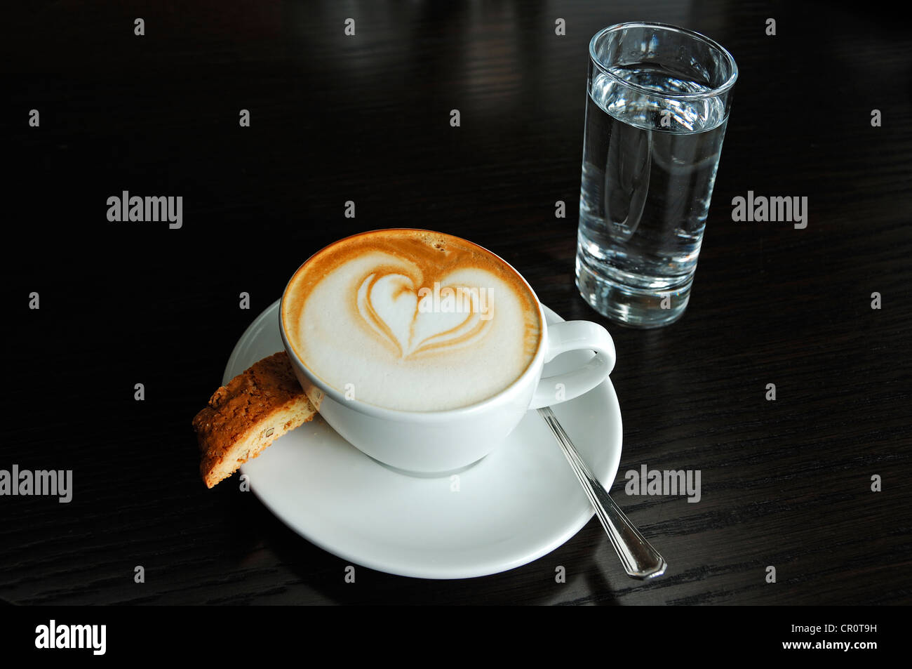 Tasse de cappuccino décoré d'un coeur, une biscotte et un verre d'eau sur un tableau noir Banque D'Images