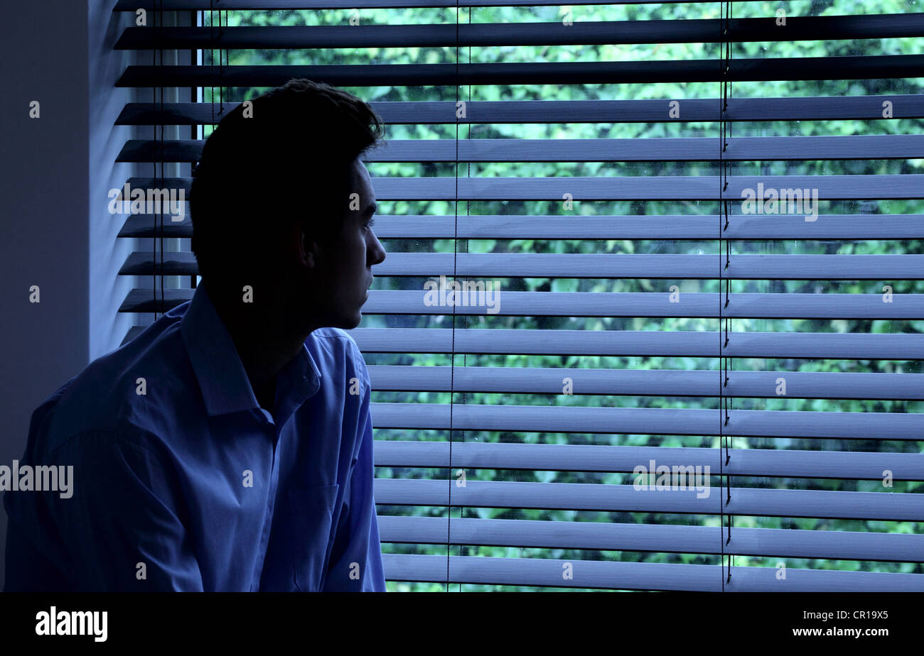 Jeune homme assis dans une pièce sombre à la recherche à travers une fenêtre aveugle. Banque D'Images