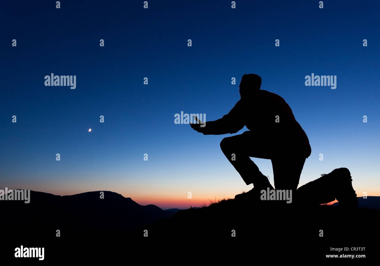 Homme priant pour le pardon sur le sommet d'une montagne au coucher du soleil avec la lune dans le ciel. Banque D'Images Homme priant pour le pardon sur le sommet d'une montagne au coucher du soleil avec la lune dans le ciel. Banque D'Images