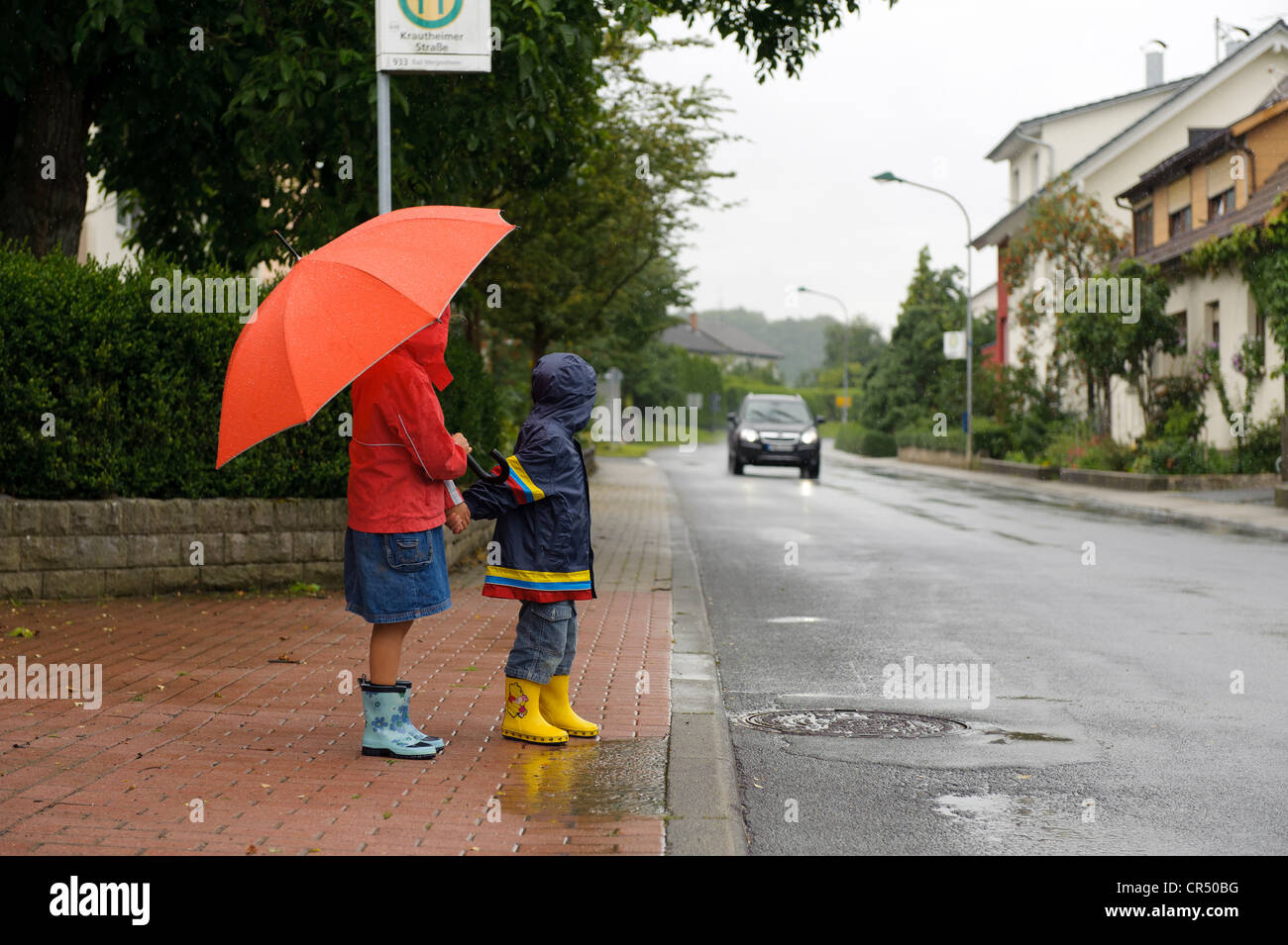 Deux enfants, 3 et 7 ans, en attendant de traverser la rue sous la pluie, bien qu'une voiture approche, Assamstadt, Bade-Wurtemberg Banque D'Images