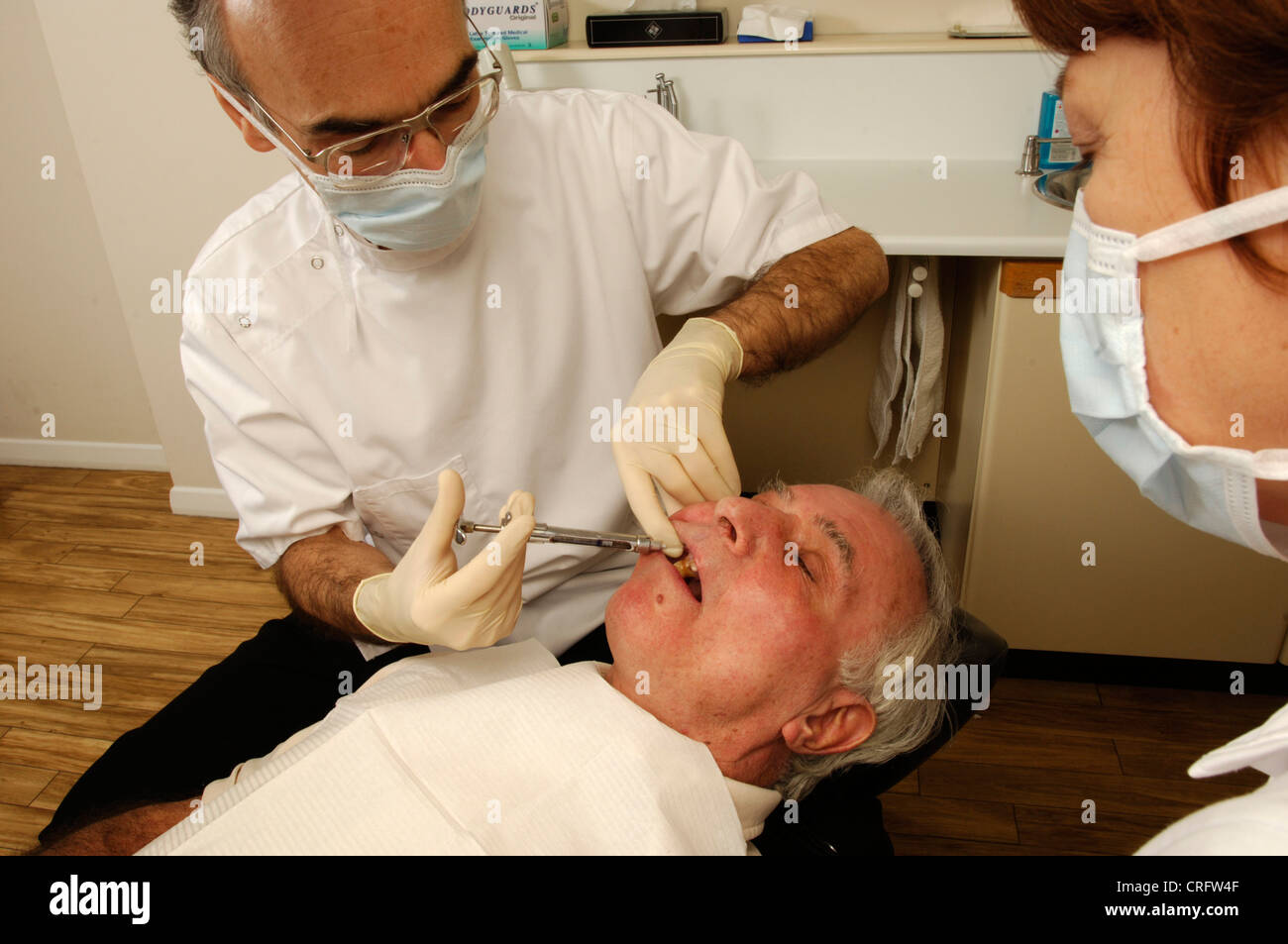 Vu par son adjoint, un dentiste injecte un anesthésique local anti douleur dans une gomme du patient pour engourdir la région avant de commencer une procédure dentaire. Banque D'Images