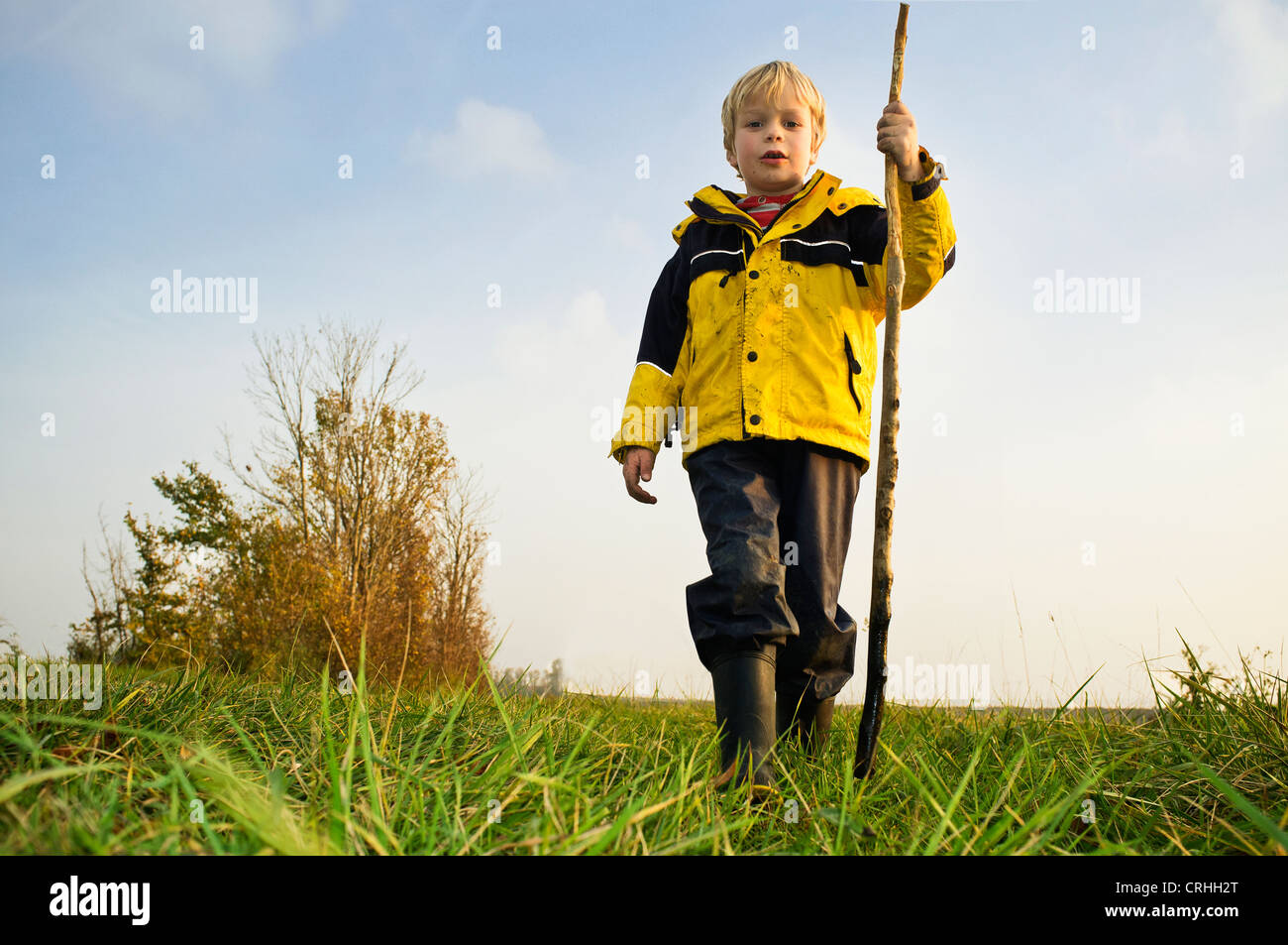 Jeune enfant, portant un manteau de pluie jaune et bottes, posant dans un paysage rural avec un bâton Banque D'Images
