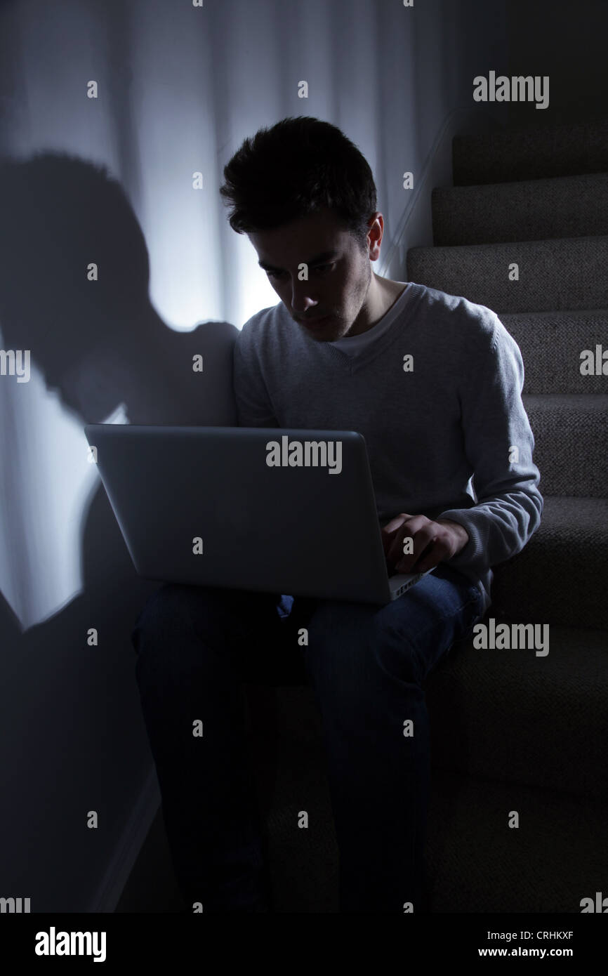 Teenage male en utilisant son ordinateur portable, assis dans l'escalier sombre.. Banque D'Images
