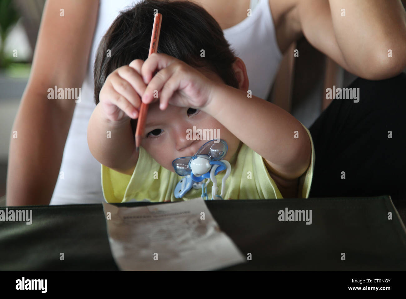 C'est une photo d'un jeune garçon chinois bébé qui essaie d'écrire avec un stylo sur une feuille de papier. Il a une sucette dans la bouche. Jour Banque D'Images